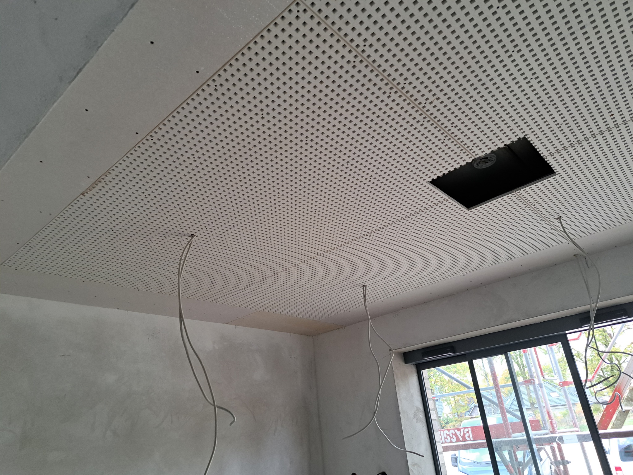 Montaż perforowanej płyty karton gips na suficie z otworem na oświetlenie, widoczne kable i surowe ściany. W tle okno z widokiem na zewnątrz.