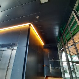 Mieszko Zieliński - Nowoczesne oświetlenie LED wzdłuż sufitu i ściany, z widocznym wejściem do windy i fragmentem przeszklonej ściany z metalową konstrukcją w tle.