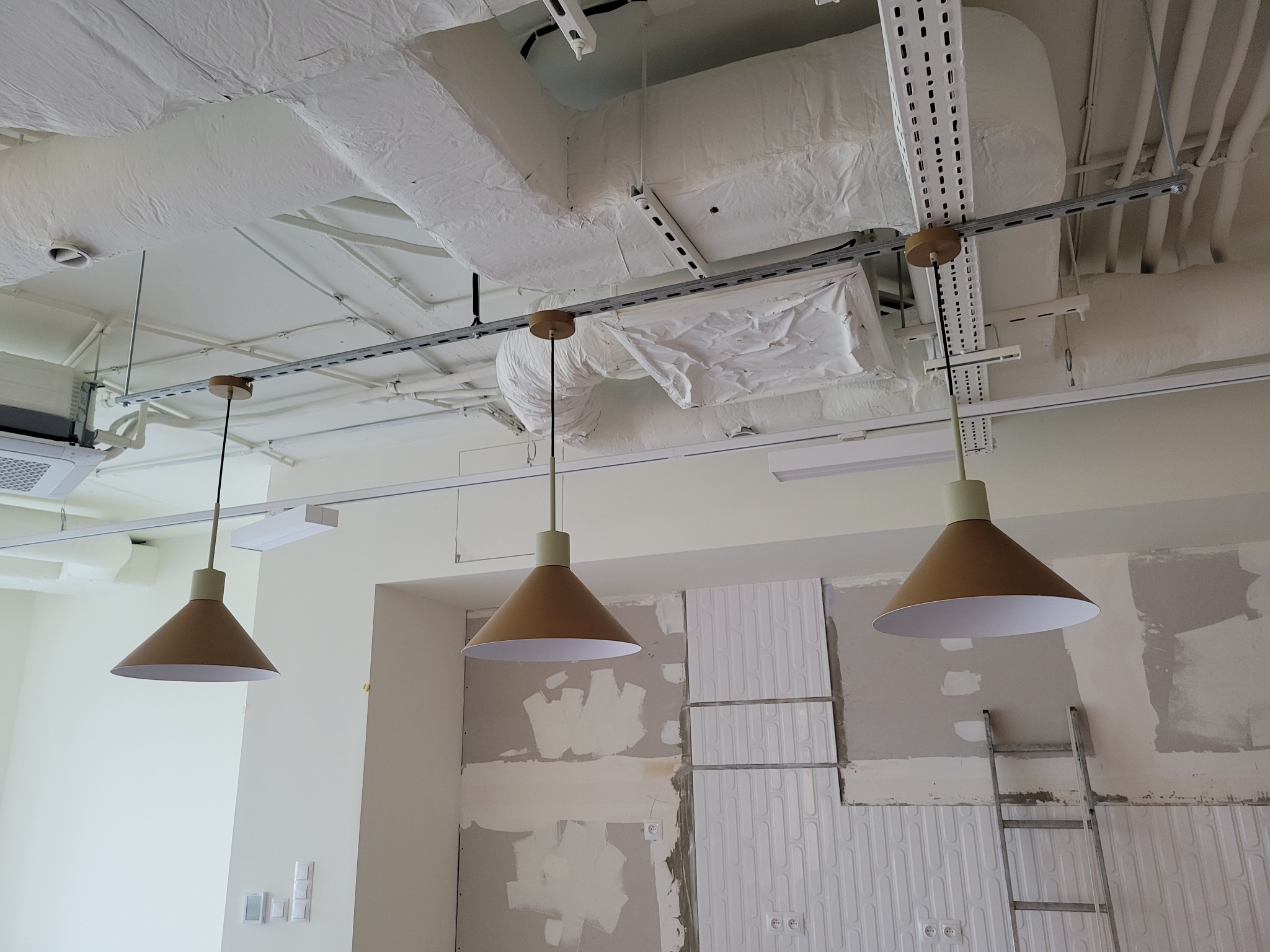 Trzy lampy stożkowe zawieszone na szynie w jasnym wnętrzu w trakcie remontu, z widocznymi instalacjami pod sufitem i fragmentami ścian przygotowanych do wykończenia.