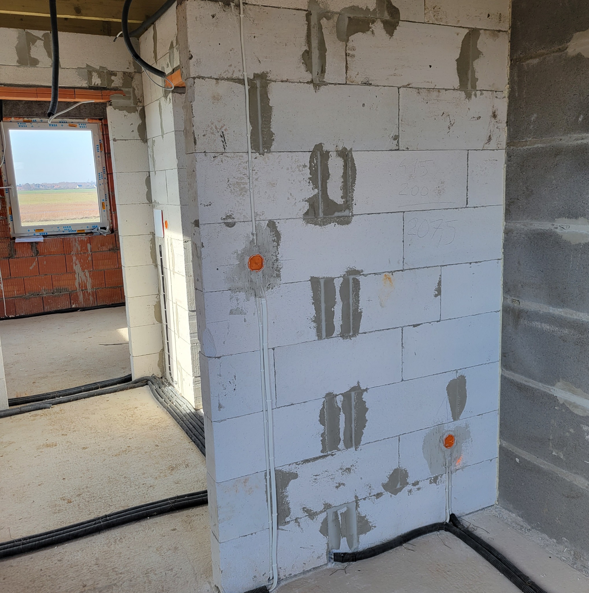 Wnętrze w trakcie prac: białe ściany z bloczków, widoczne peszle elektryczne w podłodze i przewody w ścianach, przygotowane pod instalacje elektryczne w nowym budynku.