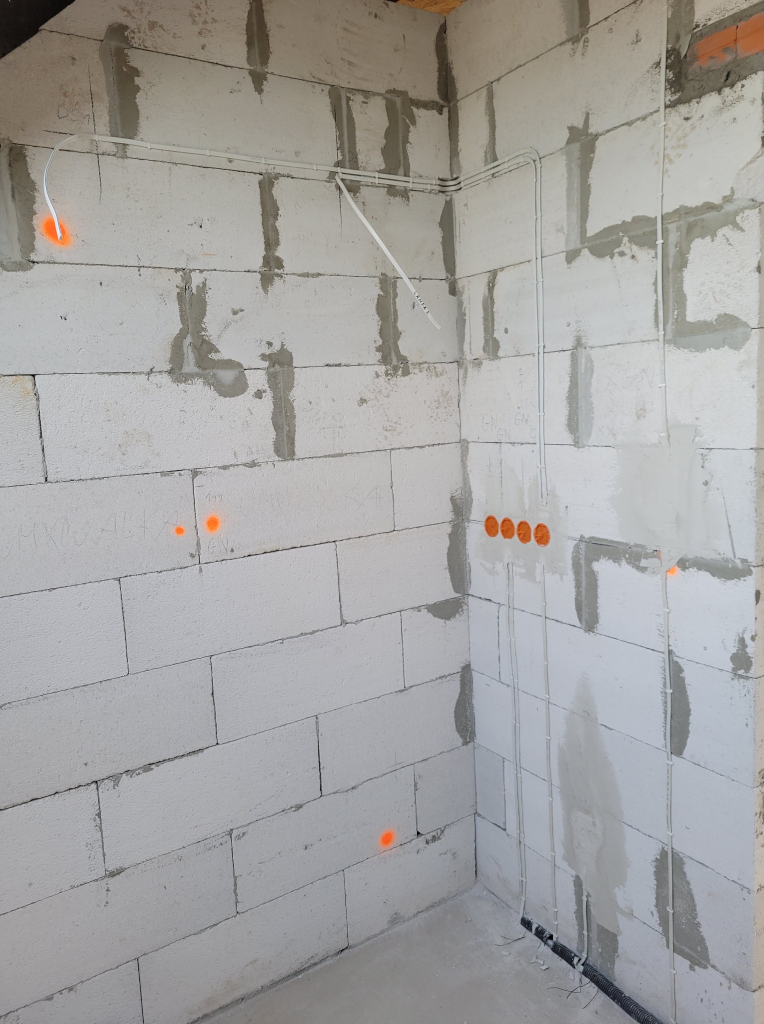 Surowe ściany z betonu komórkowego z rozprowadzoną instalacją elektryczną w białych rurkach. Widoczne pomarańczowe oznaczenia punktów elektrycznych.