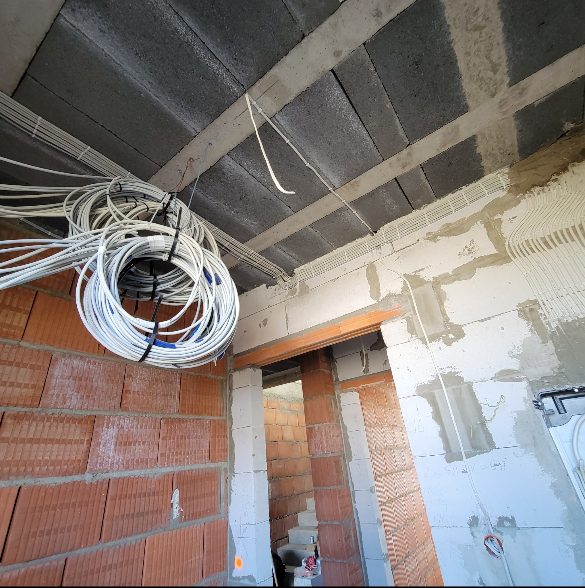 Kable elektryczne zebrane w pęczki, prowadzone pod sufitem budynku w trakcie budowy. Widoczne ceglane ściany i surowy betonowy strop.