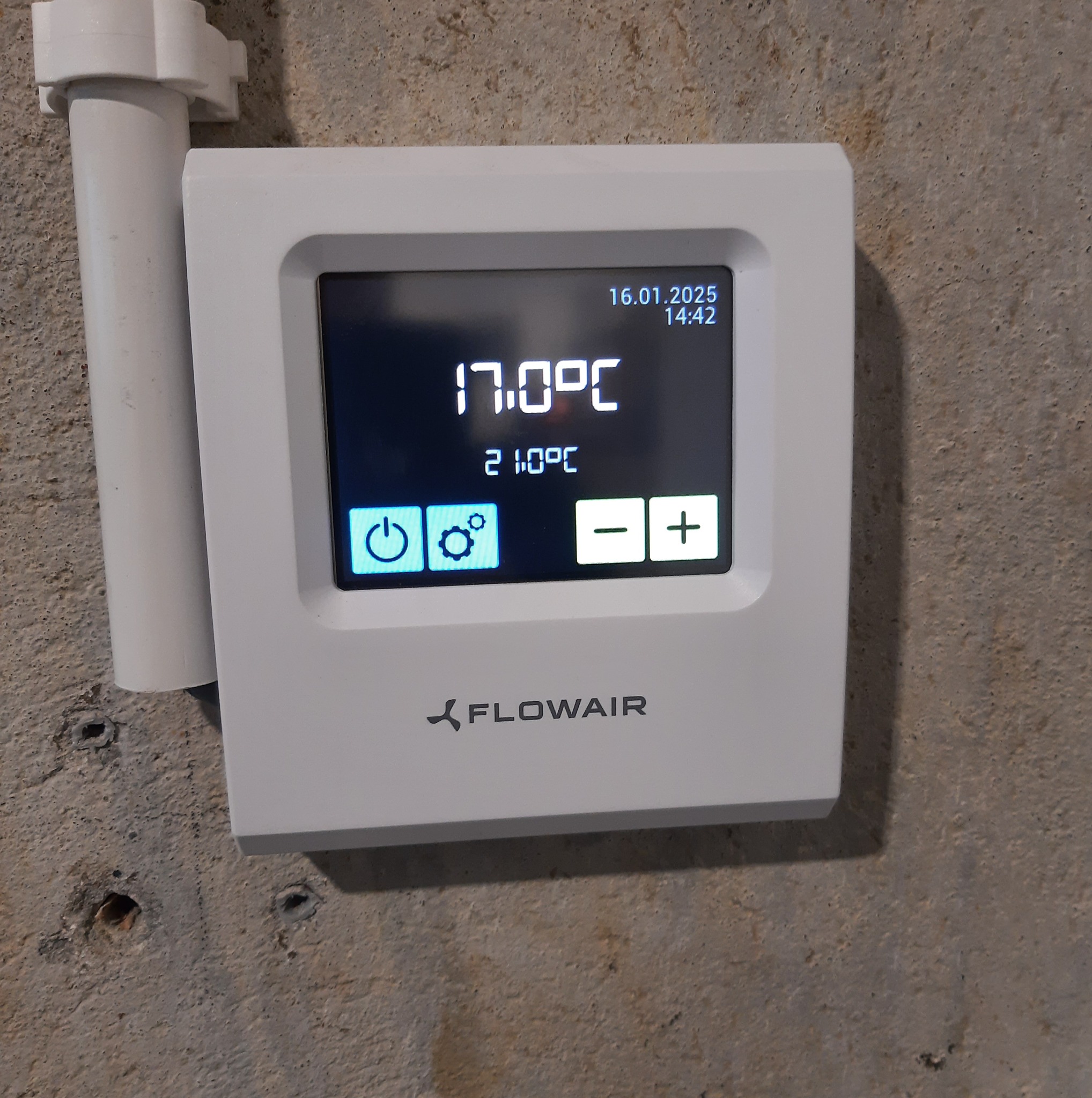 Biały, cyfrowy termostat Flowair zamontowany na szarej ścianie, wyświetlający temperaturę 17 stopni Celsjusza. Widoczny fragment rury instalacyjnej.