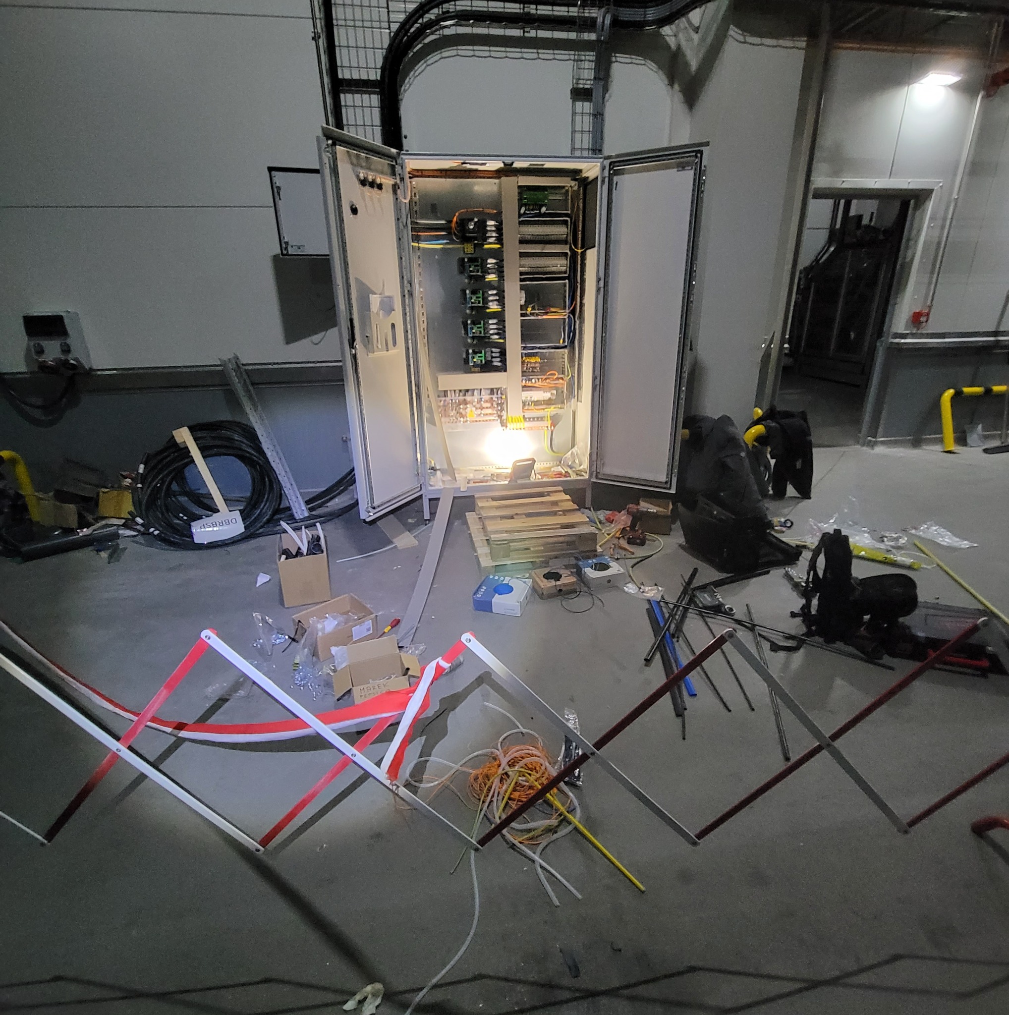 Otwarta szafa elektryczna z widocznymi kablami i elementami sterującymi wewnątrz, otoczona narzędziami i zabezpieczeniami na podłodze w hali przemysłowej.