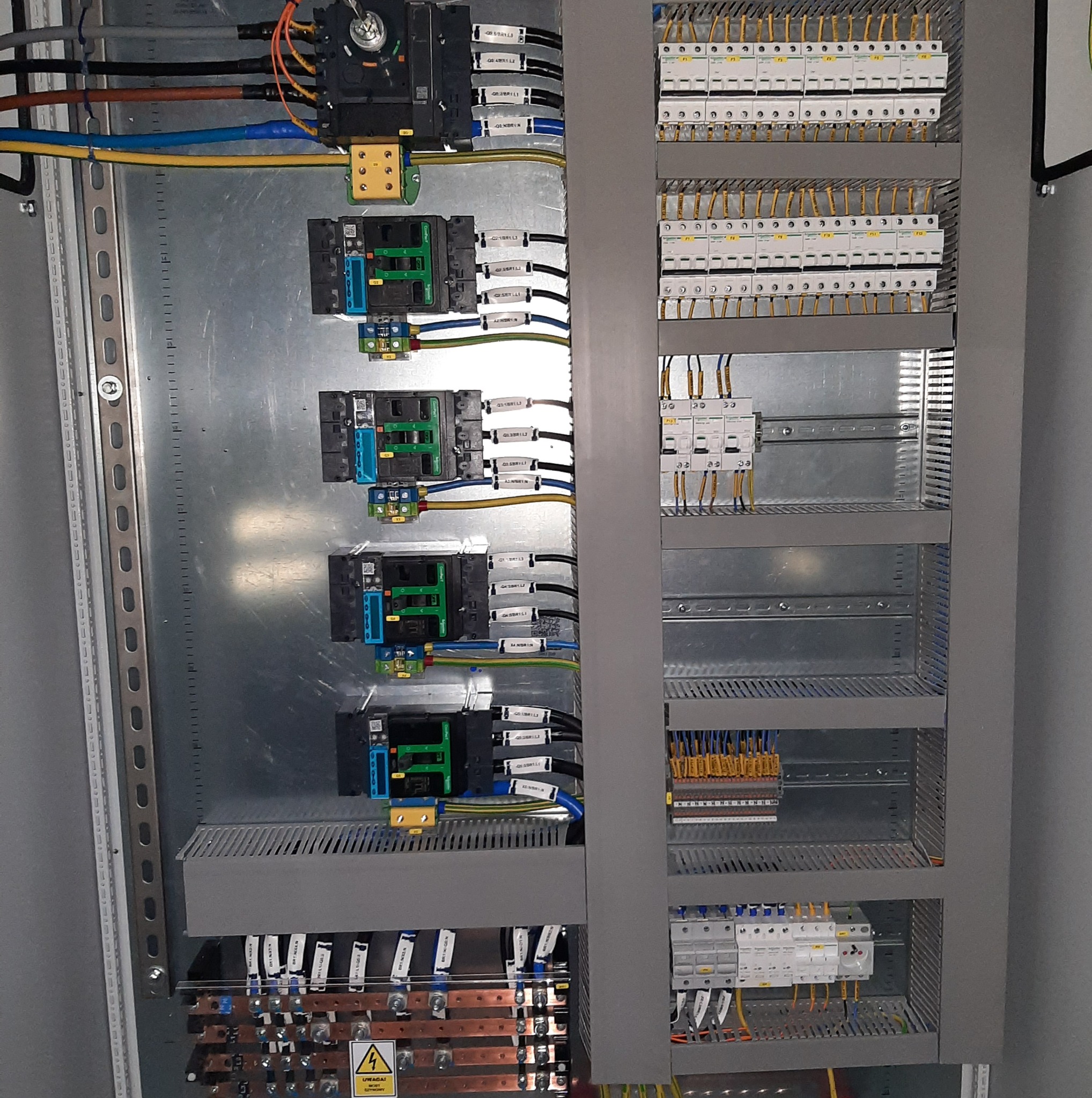 Szara szafa sterownicza z zainstalowanymi modułami, bezpiecznikami i okablowaniem. Widoczne listwy zaciskowe, przewody w różnych kolorach i ostrzeżenie o napięciu.