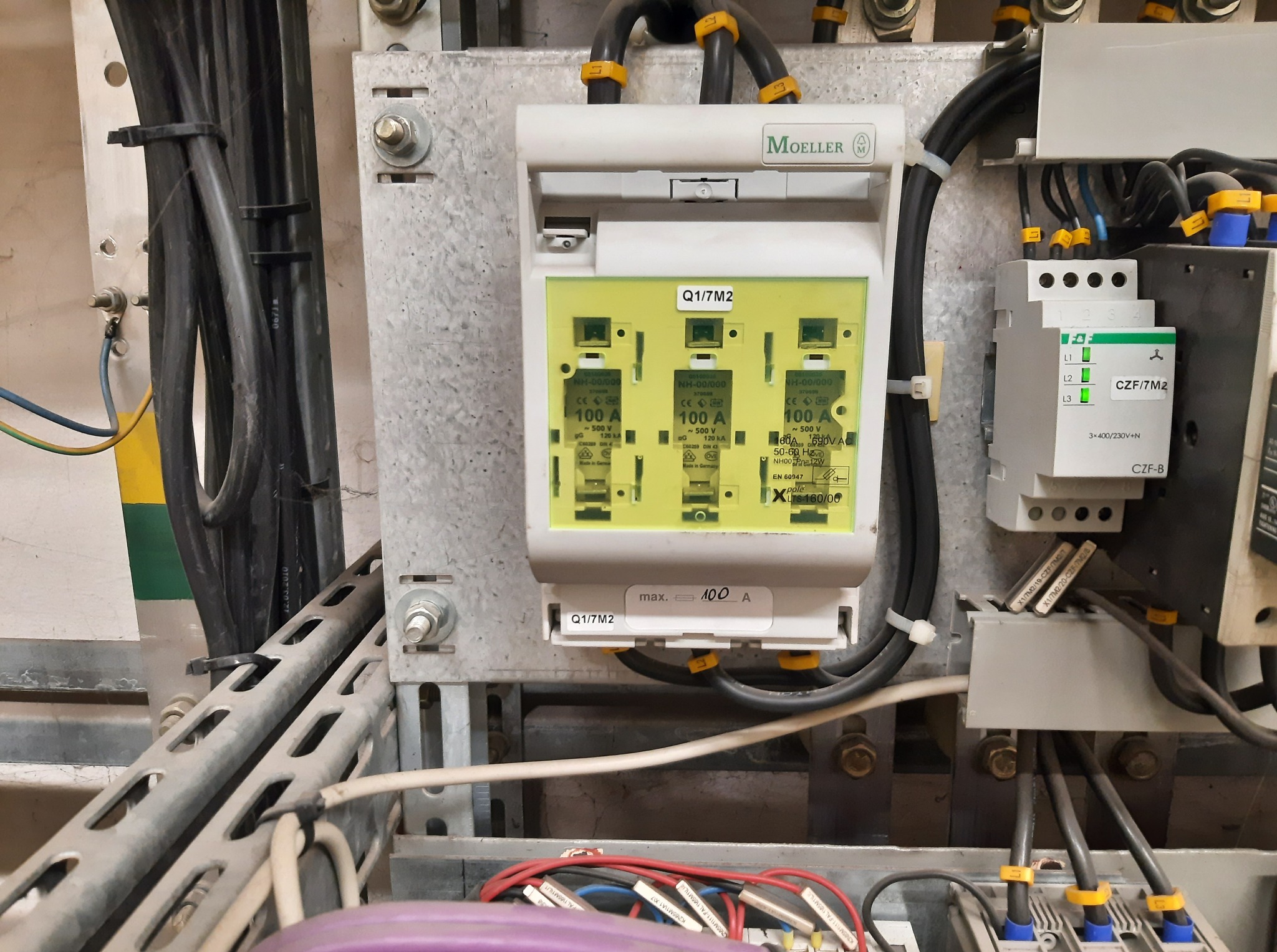 Tablica rozdzielcza z bezpiecznikami Moeller 100A i okablowaniem w metalowej obudowie. Widoczne przewody z oznaczeniami i elementy sterownicze.