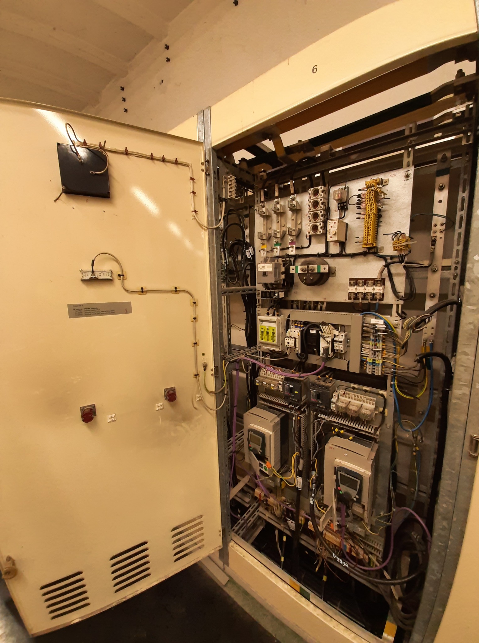 Otwarta szafa sterownicza z widocznymi elementami elektrycznymi, przewodami i urządzeniami. Panel w kolorze beżowym, widoczne bezpieczniki, przekaźniki i okablowanie.