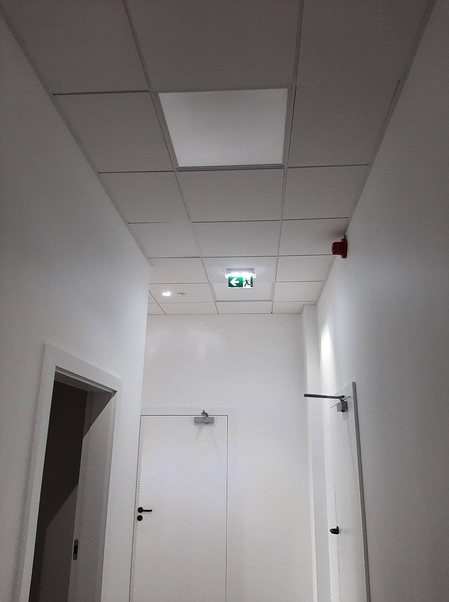 Korytarz biurowy z podwieszanym sufitem, oświetleniem LED i znakiem ewakuacyjnym. Widoczny czujnik dymu i drzwi. Minimalistyczny design, surowe wykończenie.