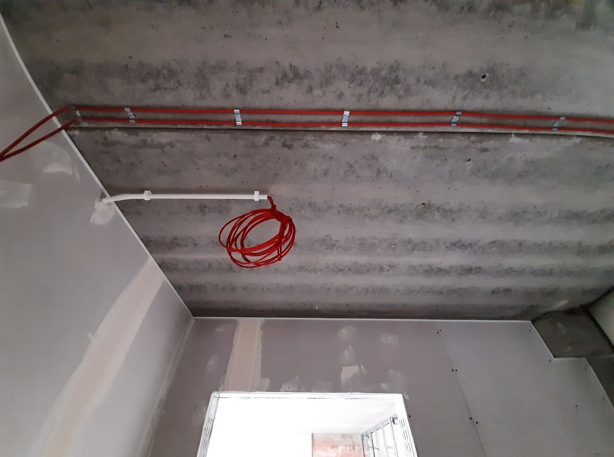 Czerwone przewody elektryczne zamocowane do sufitu w surowym wnętrzu z płyt gipsowych. Widoczne uchwyty mocujące i zwisający kabel.