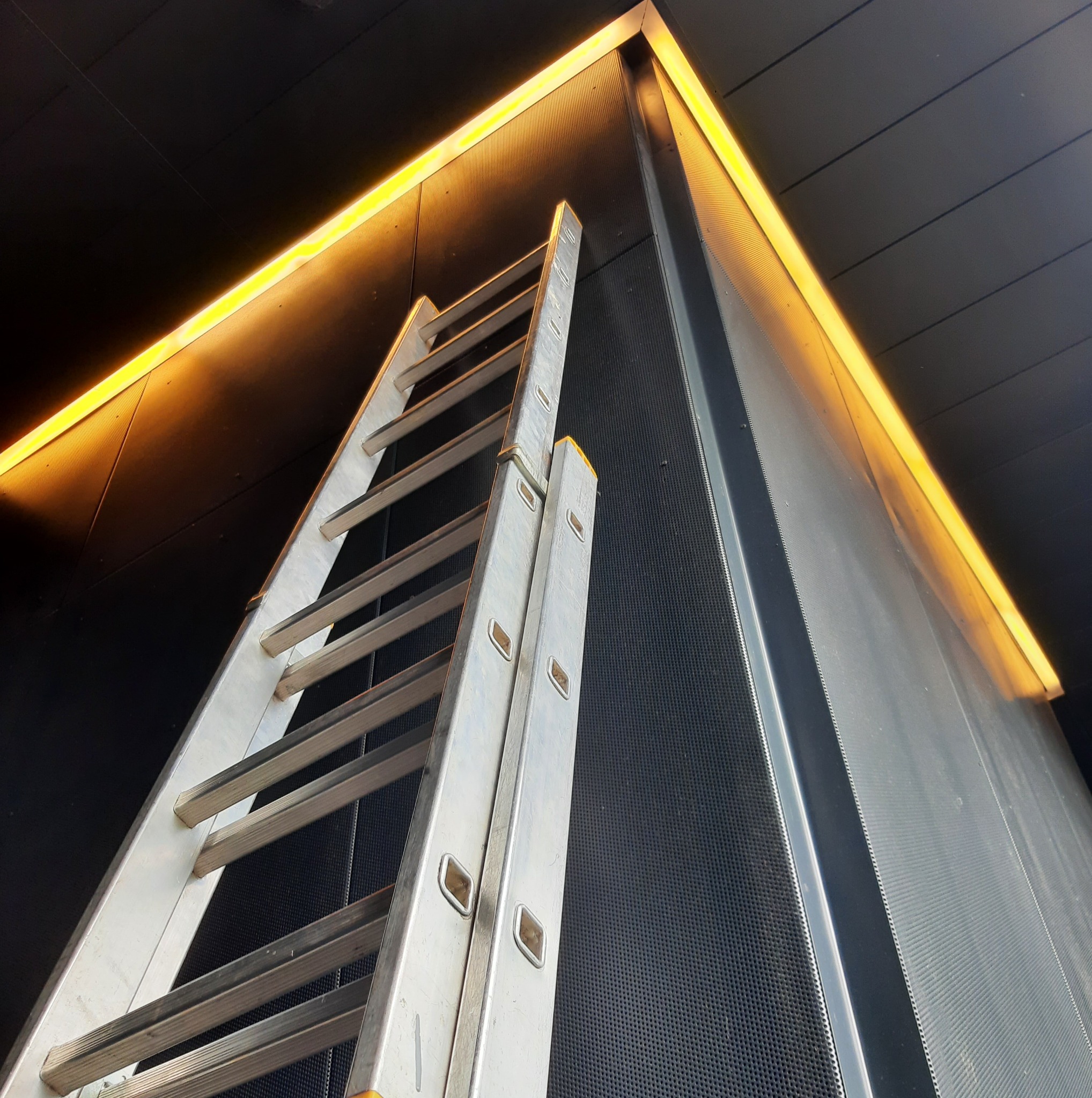 Aluminiowa drabina oparta o czarną ścianę z oświetleniem LED wzdłuż krawędzi, perspektywa z dołu, sugerująca prace instalacyjne lub konserwacyjne.