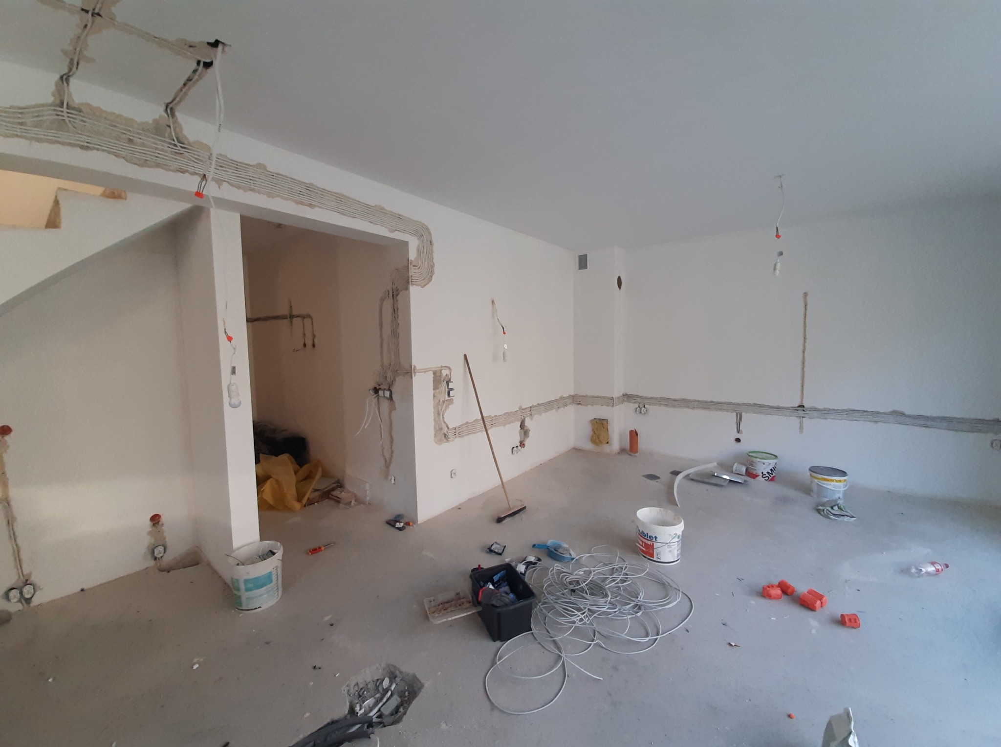 Rozległe wnętrze w trakcie prac elektrycznych: widoczne kable w bruzdach ścian i sufitu, narzędzia, puszki z farbą i elementy instalacji na surowym podłożu.