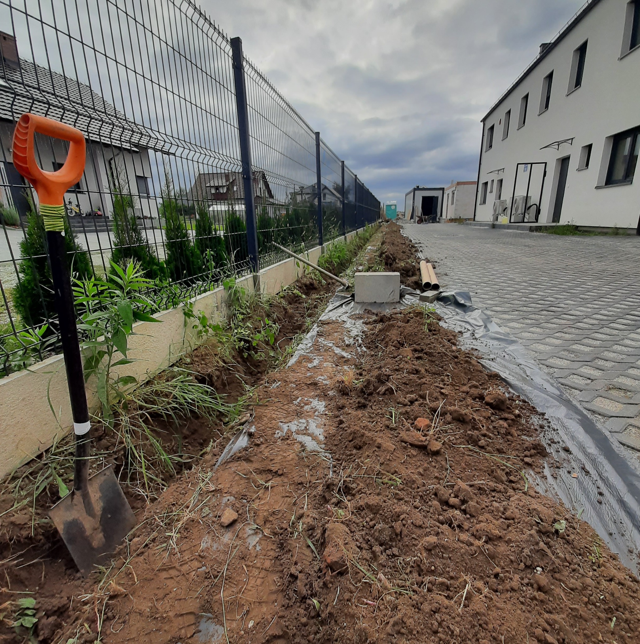 Przygotowanie terenu pod instalacje wzdłuż ogrodzenia panelowego: wykopany rów, widoczna folia, rury i szpadel z pomarańczową rączką. Budynek w tle.