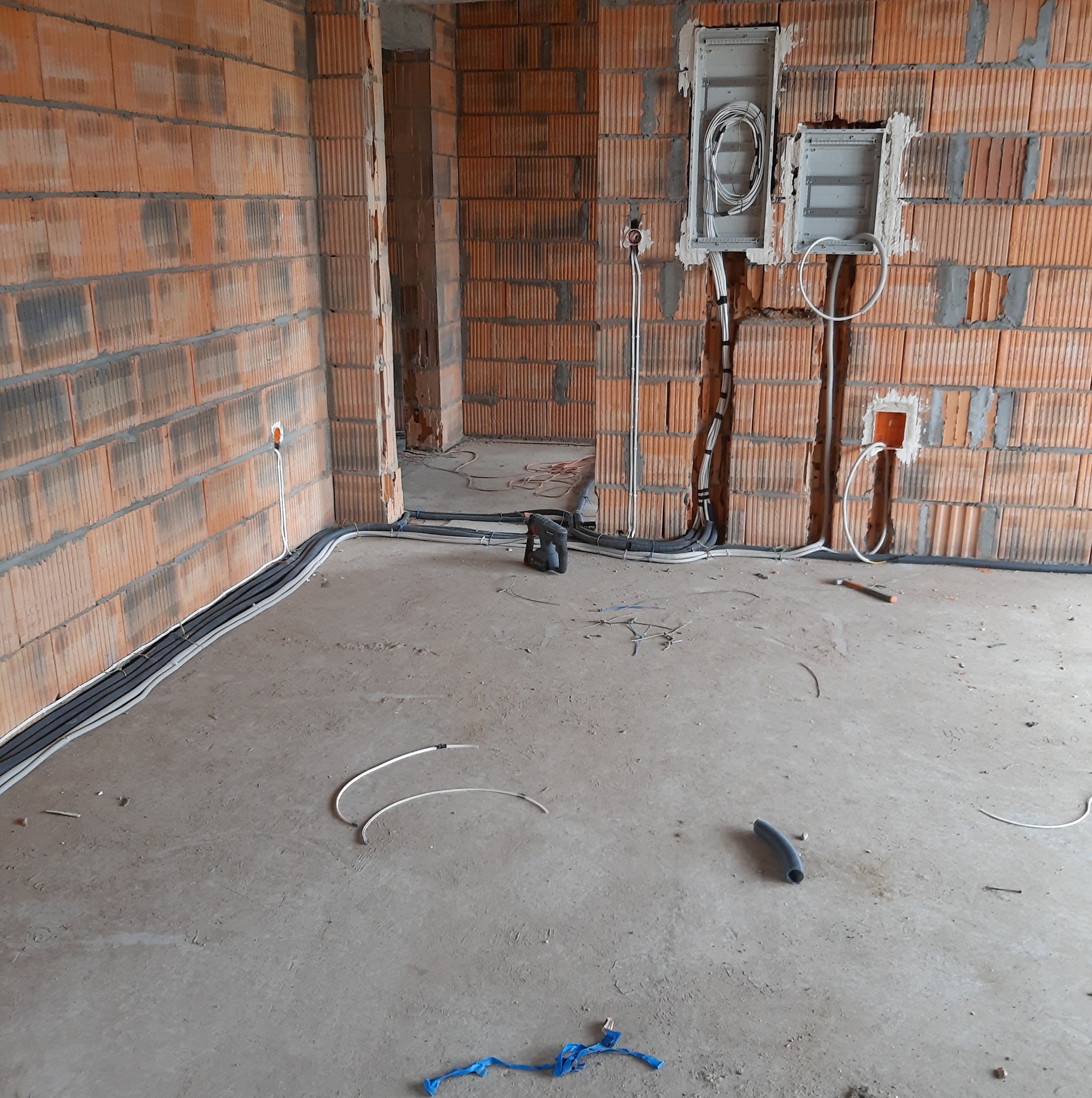 Wnętrze w trakcie prac: kable elektryczne ułożone wzdłuż ścian z czerwonej cegły, puszki podtynkowe z przewodami. Widoczne narzędzia i elementy instalacji.