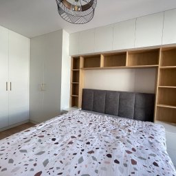 DJ Meble - Białe szafy zintegrowane z wezgłowiem łóżka i otwartymi półkami z jasnego drewna. Nad łóżkiem designerska lampa. Całość tworzy spójną zabudowę sypialnianą.