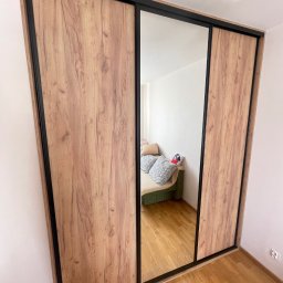 DJ Meble - Szafa wnękowa z przesuwnymi drzwiami, okleina drewnopodobna, czarne obramowanie. W lustrze odbicie sofy z poduszkami. Widok z dołu, pod kątem.