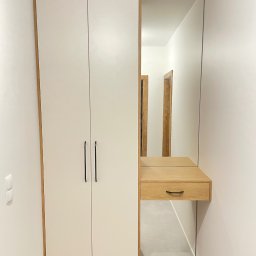 DJ Meble - Biała szafa z lustrem i drewnianą konsolą w wąskim korytarzu. Minimalistyczny design, czarne uchwyty, subtelne oświetlenie. Widoczne drzwi w tle.