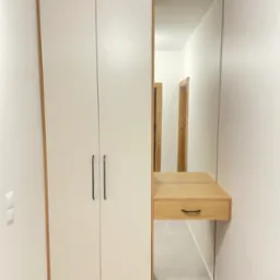 Biała szafa z lustrem i drewnianą konsolą w wąskim korytarzu. Minimalistyczny design, czarne uchwyty, subtelne oświetlenie. Widoczne drzwi w tle.