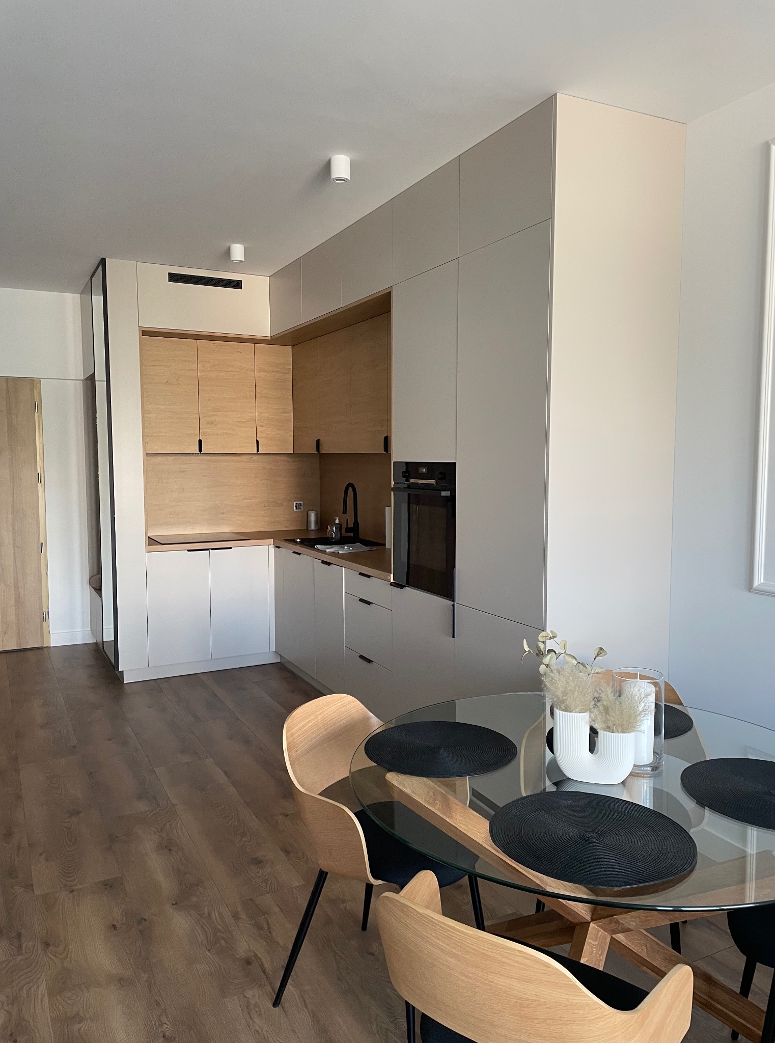 Nowoczesna kuchnia z drewnianymi akcentami i jasnymi frontami, okrągły stół ze szklanym blatem i dekoracjami. Minimalistyczny design, czarne dodatki i drewniana podłoga.