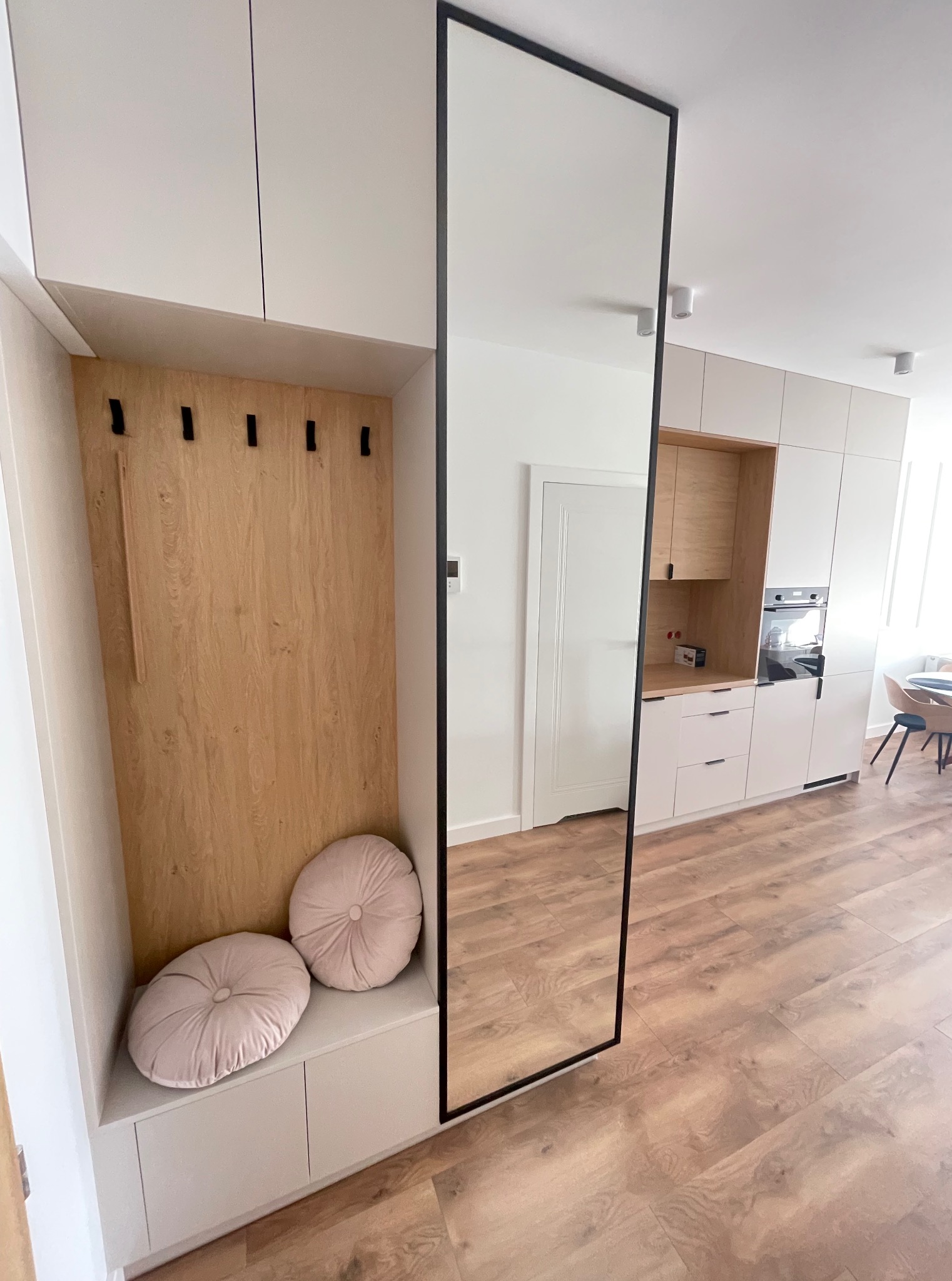 Biała zabudowa przedpokoju z siedziskiem, wnęką z wieszakami i dużym lustrem w czarnej ramie. W tle zabudowa kuchenna w podobnym stylu.