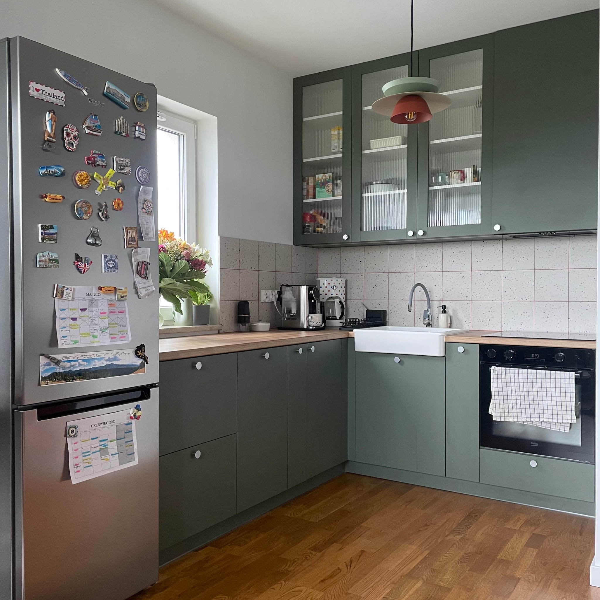 Kuchenna zabudowa w odcieniach zieleni z ryflowanymi szybami w szafkach wiszących. Na lodówce magnesy z podróży i kalendarz. Drewniany blat i podłoga.