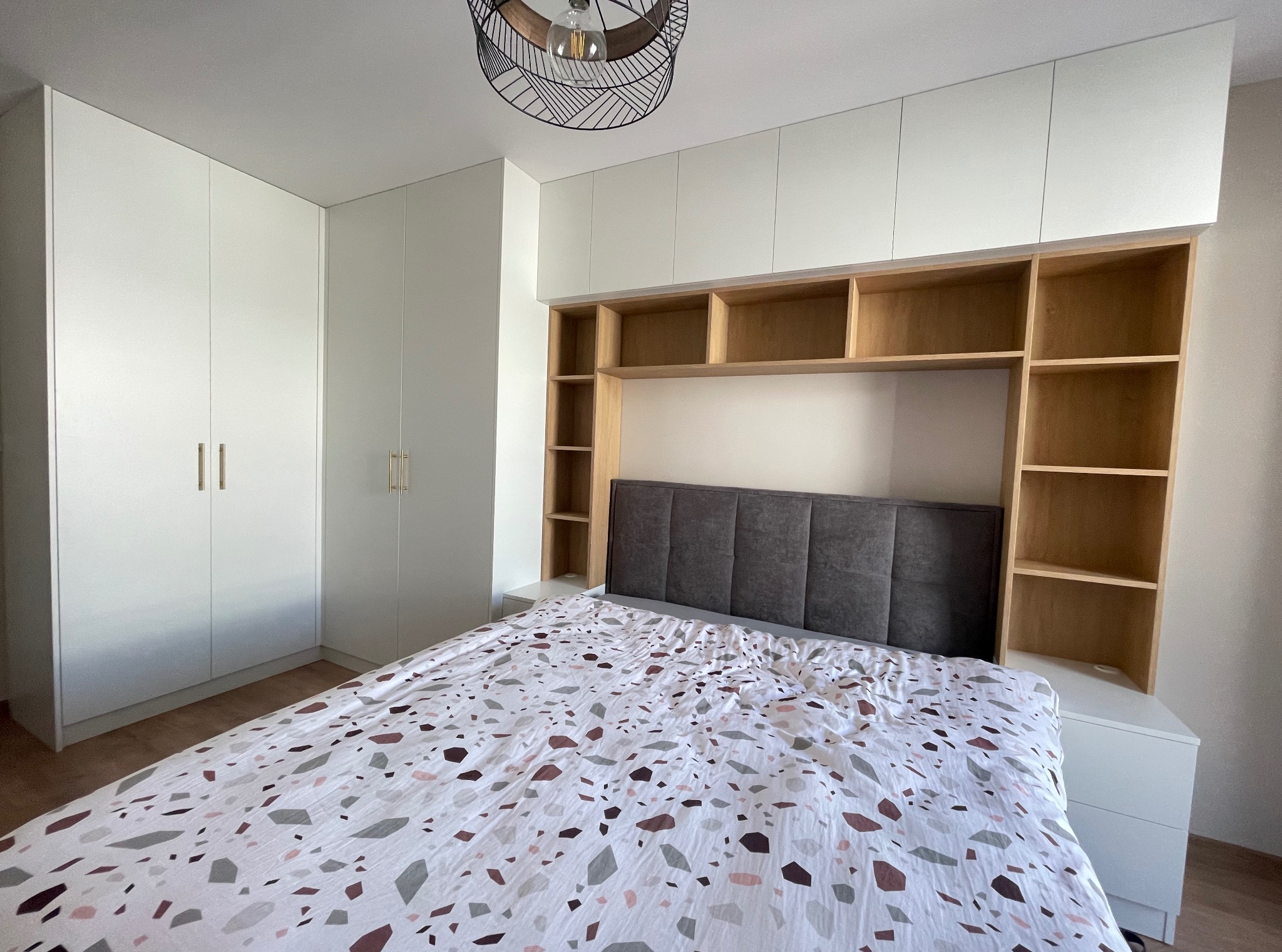 Białe szafy zintegrowane z wezgłowiem łóżka i otwartymi półkami z jasnego drewna. Nad łóżkiem designerska lampa. Całość tworzy spójną zabudowę sypialnianą.