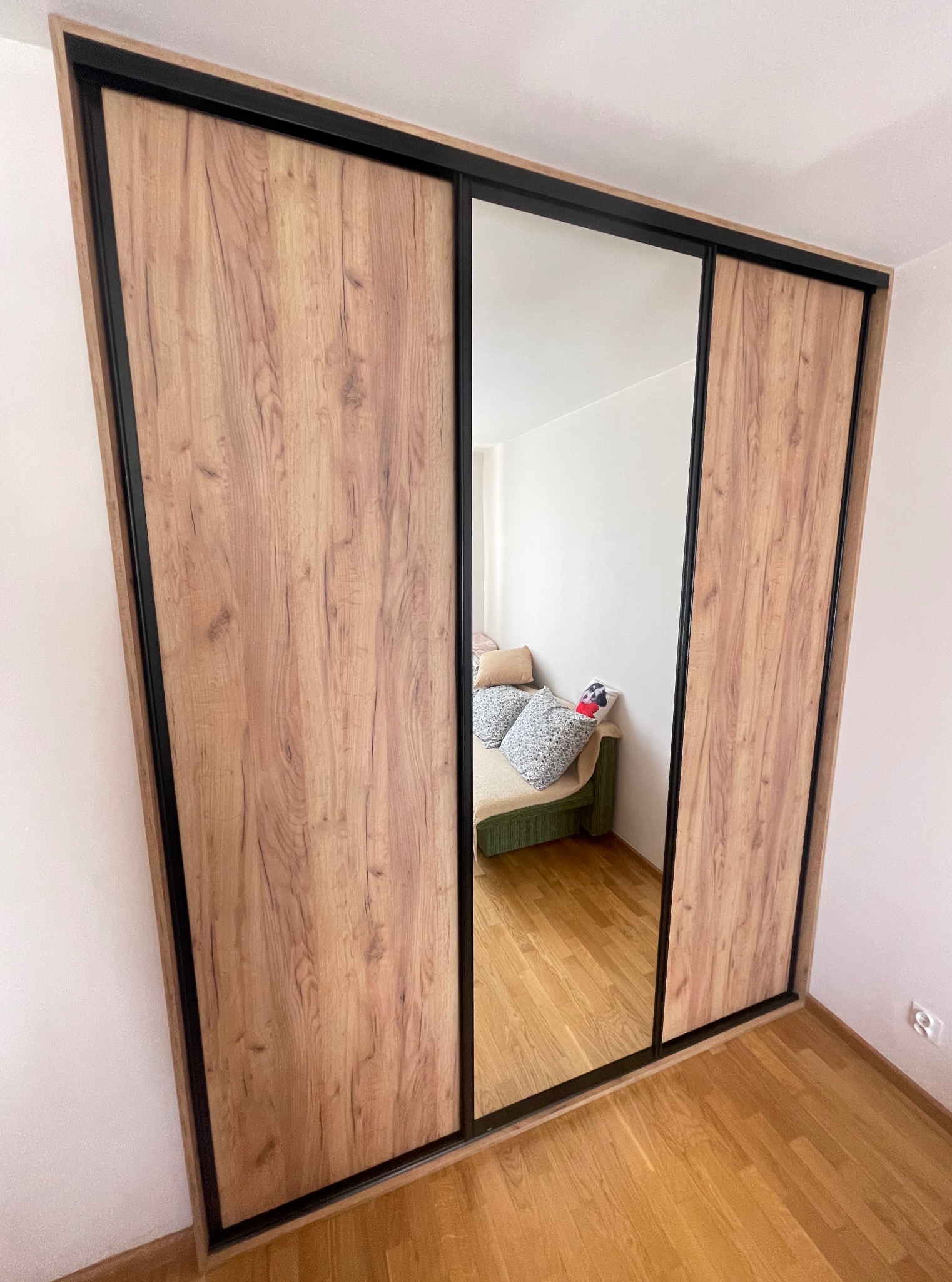 Szafa wnękowa z przesuwnymi drzwiami, okleina drewnopodobna, czarne obramowanie. W lustrze odbicie sofy z poduszkami. Widok z dołu, pod kątem.