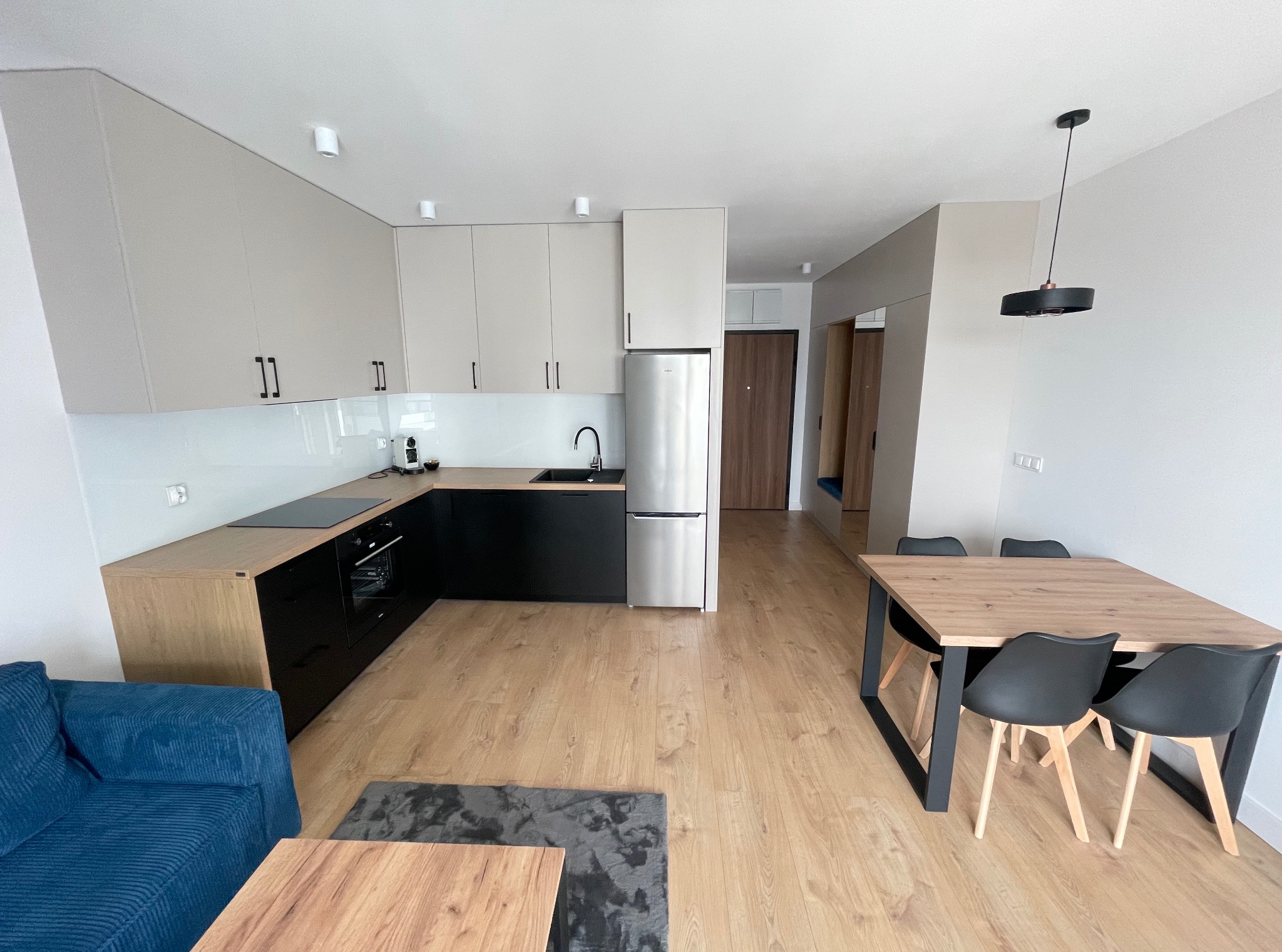 Jasna, nowoczesna kuchnia z czarnymi szafkami dolnymi i beżowymi górnymi. Widoczny stół z krzesłami i fragment niebieskiej sofy. Minimalistyczny design.