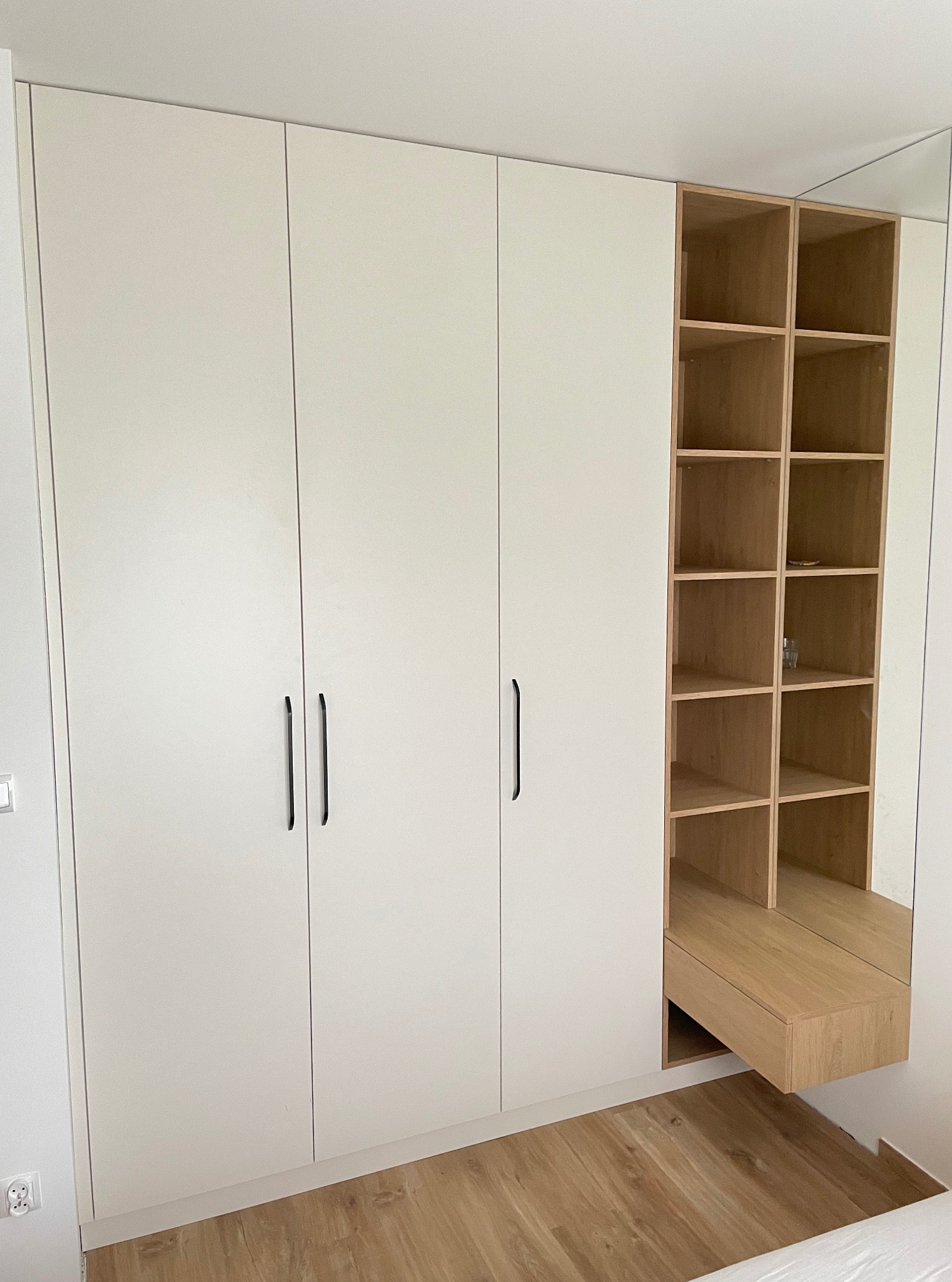 Biała szafa wnękowa z czarnymi uchwytami i drewnianym regałem z lustrem. Minimalistyczny design, jasne wnętrze, parkiet na podłodze.