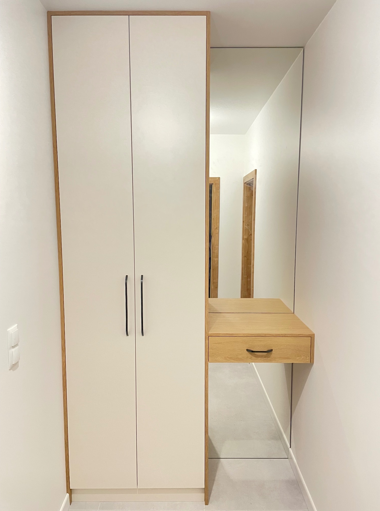 Biała szafa z lustrem i drewnianą konsolą w wąskim korytarzu. Minimalistyczny design, czarne uchwyty, subtelne oświetlenie. Widoczne drzwi w tle.