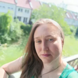 Kobieta w turkusowej bluzce na balkonie z widokiem na zielone otoczenie i budynki mieszkalne w tle. Naturalne światło, ujęcie z bliska.