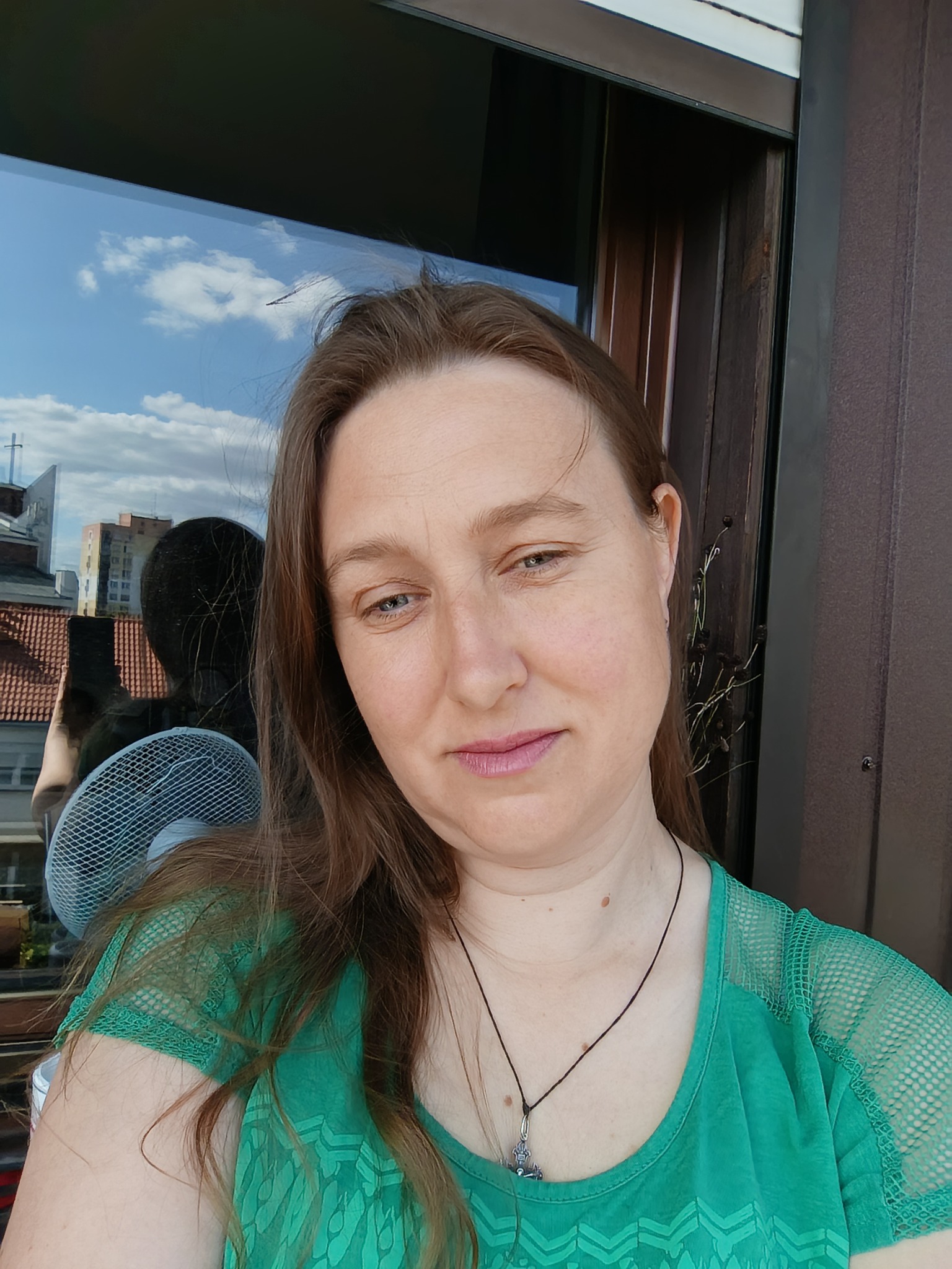 Portret kobiety w zielonej bluzce, z widokiem na miasto odbijającym się w oknie. W tle widać wentylator i brązowe panele.