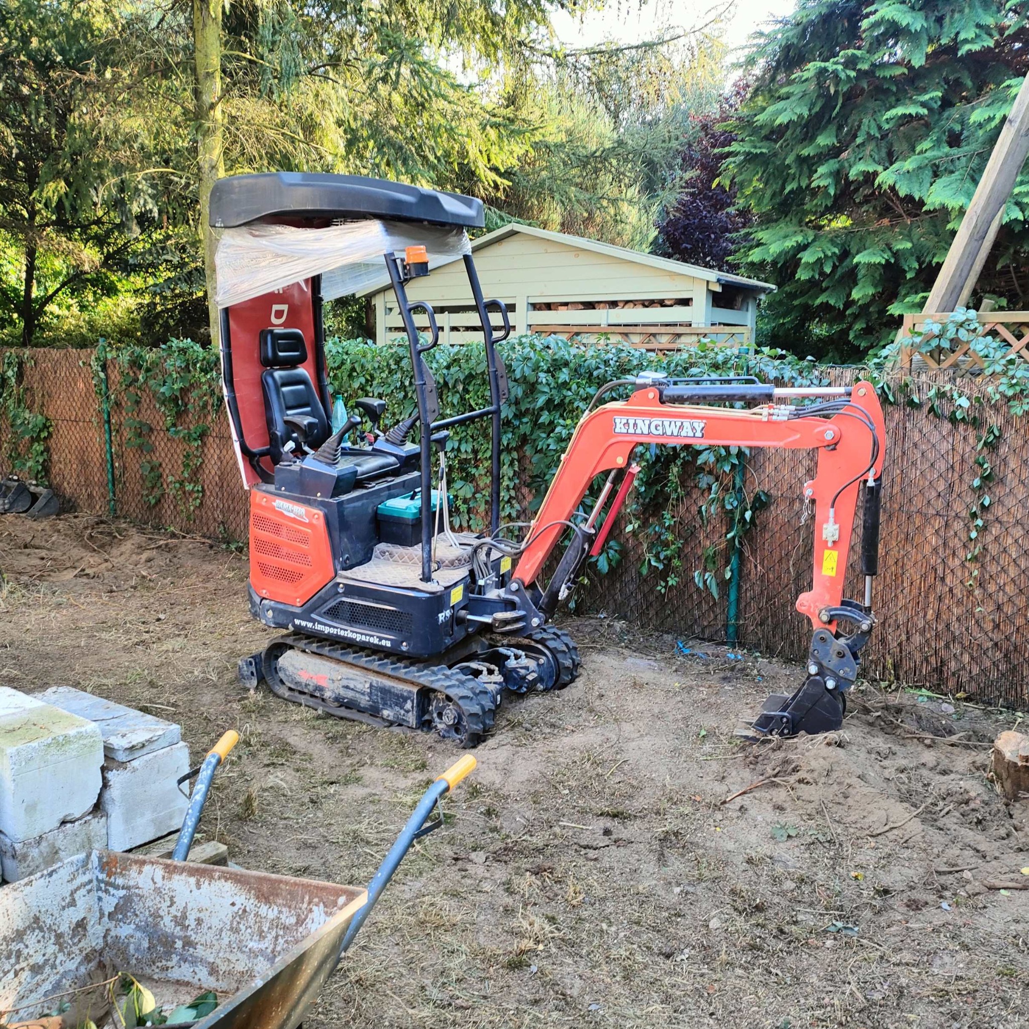 Pomarańczowa mini koparka Kingway na trawiastym terenie, z wózkiem i stertą betonowych bloków, sugerująca prace ziemne lub przygotowanie terenu pod ogród.