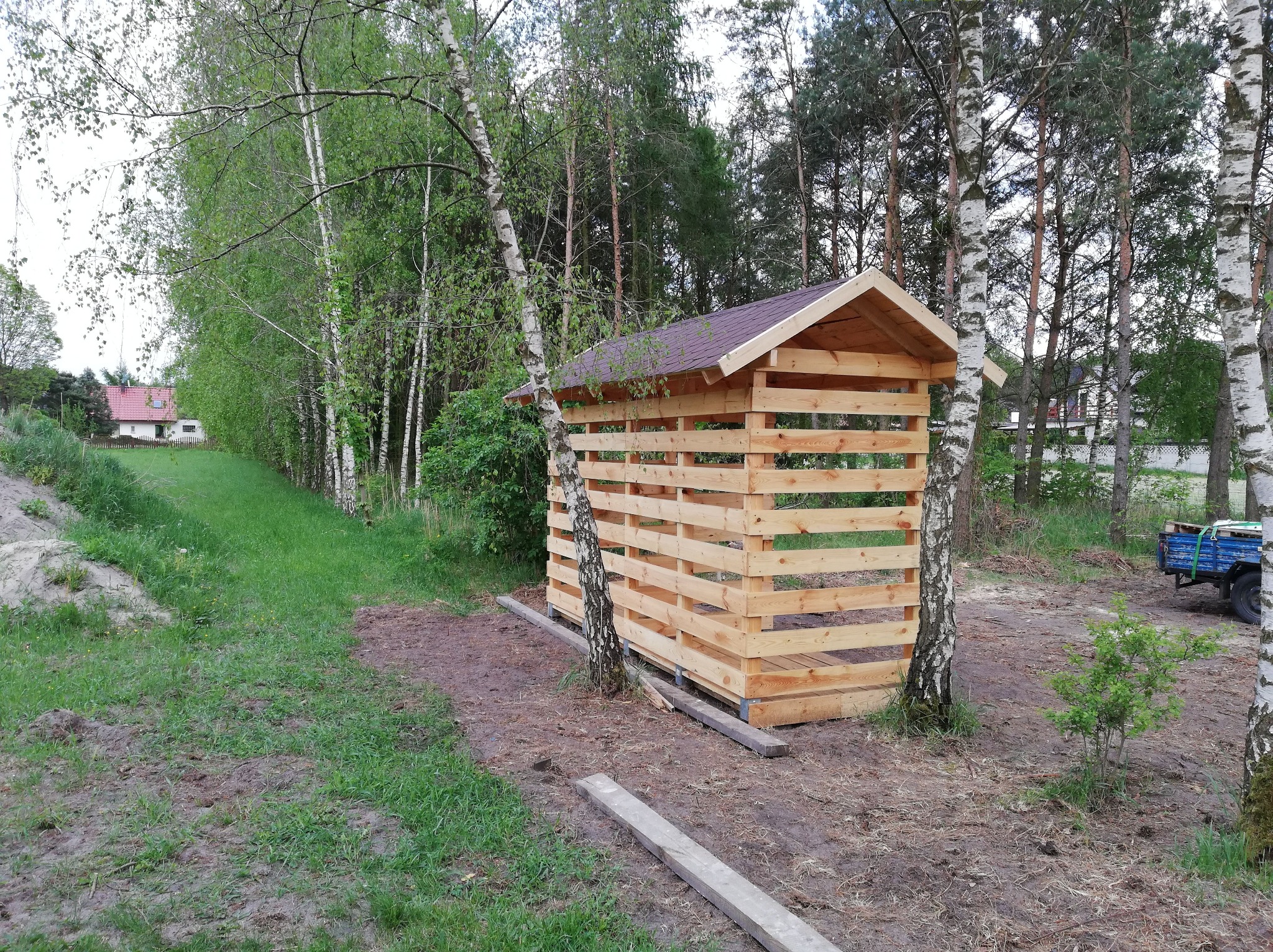 Drewniana konstrukcja z dachem w Gnieźnie, otoczona brzozami i zielenią. Widoczny fragment przyczepki i domu w oddali. Konstrukcja z belek, surowa.