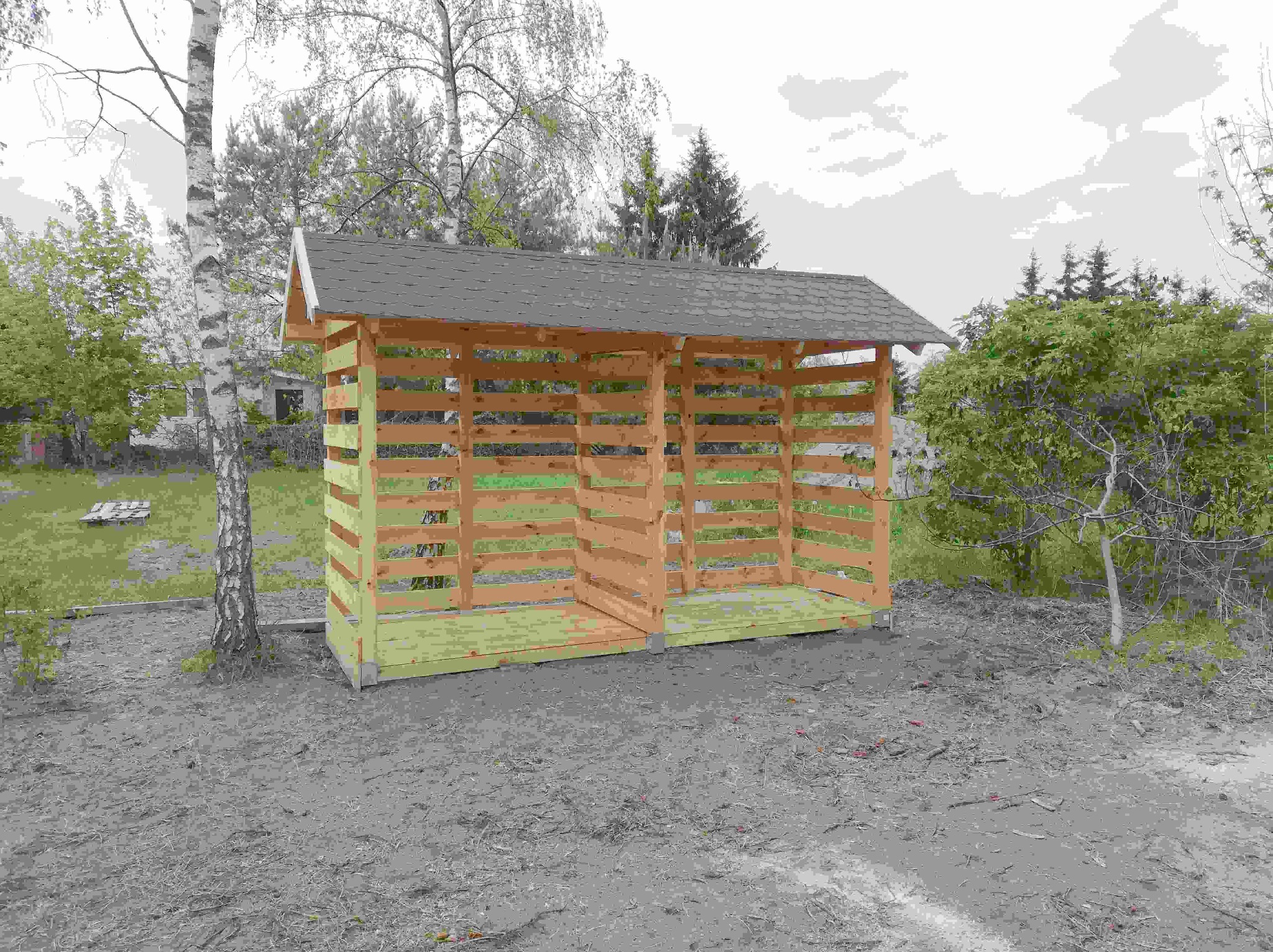 Drewniana konstrukcja z dachem w ogrodzie, możliwe przeznaczenie jako składzik na drewno lub narzędzia ogrodowe. W tle widoczne drzewa.