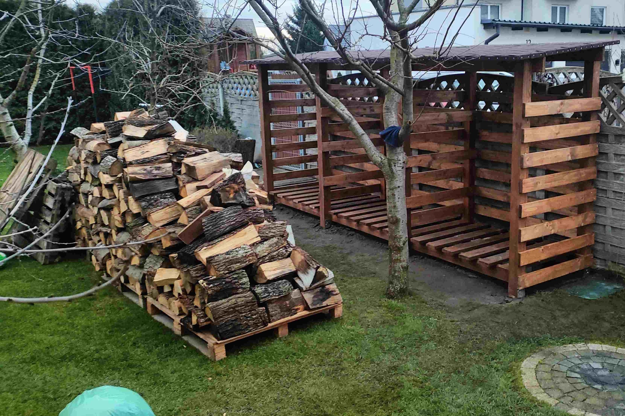 Drewniany składzik na opał z ułożonym drewnem na palecie, na trawniku. W tle widoczny płot i budynek.