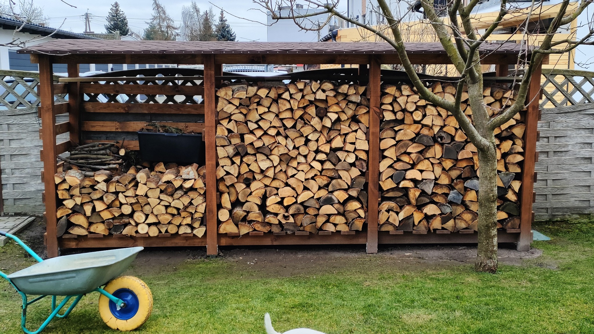 Drewniany składzik na drewno opałowe z ażurowymi ścianami, częściowo wypełniony pociętymi kawałkami drewna. Obok taczka i małe drzewo.
