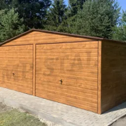 Drewnopodobny garaż blaszany z dwoma bramami, umieszczony na kostce brukowej, otoczony zielenią i drzewami w słoneczny dzień.