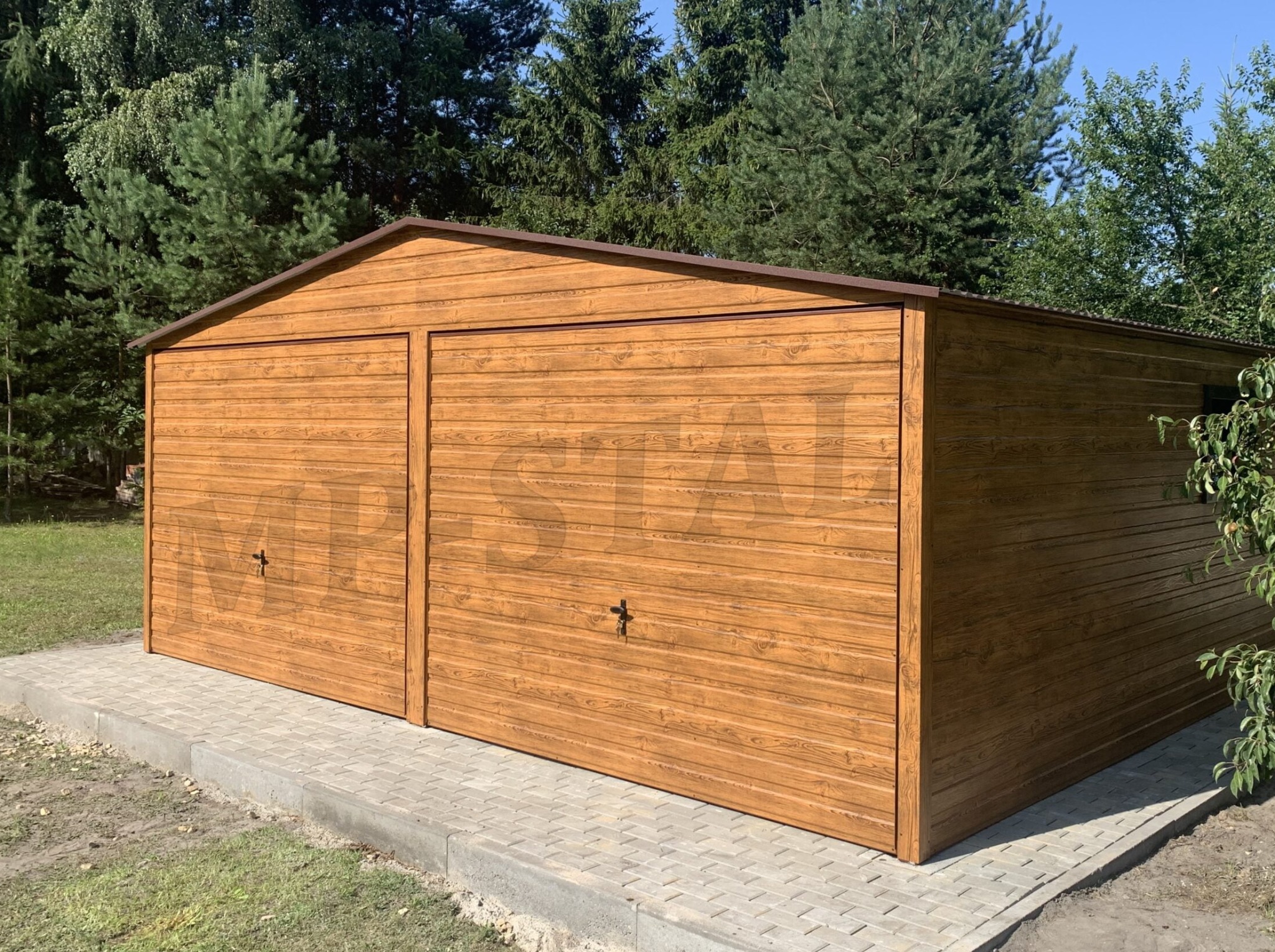 Drewnopodobny garaż blaszany z dwoma bramami, umieszczony na kostce brukowej, otoczony zielenią i drzewami w słoneczny dzień.