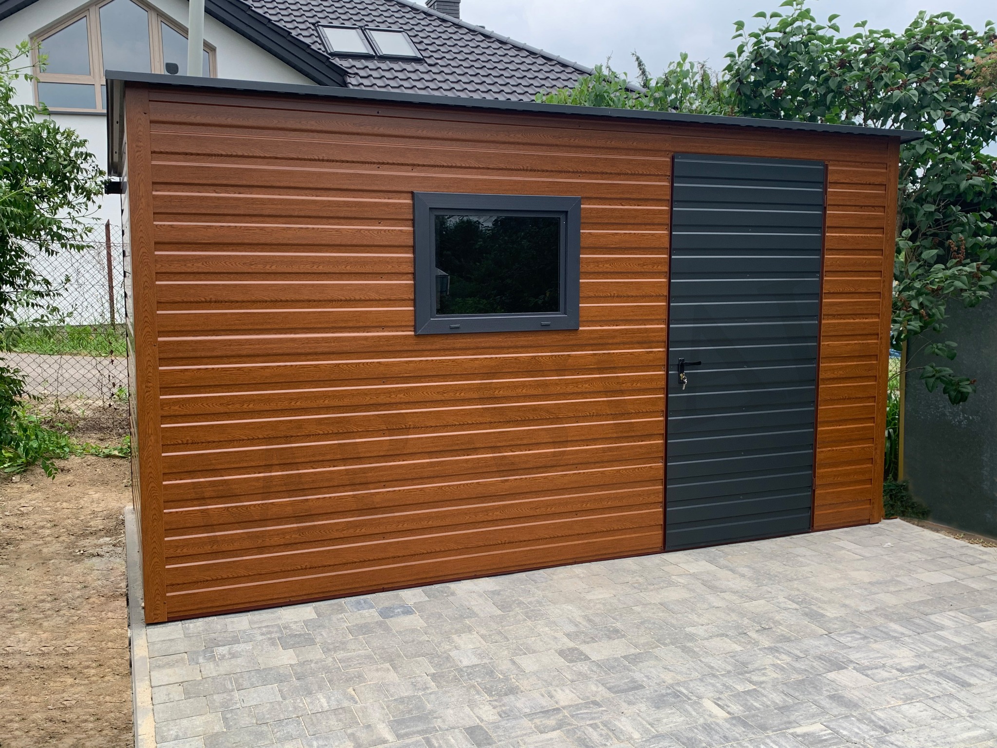 Brązowy garaż blaszany z szarą bramą i oknem, na kostce brukowej. Widok z zewnątrz, w tle dom z ciemną dachówką i zieleń. Garaż z poziomym wzorem desek.