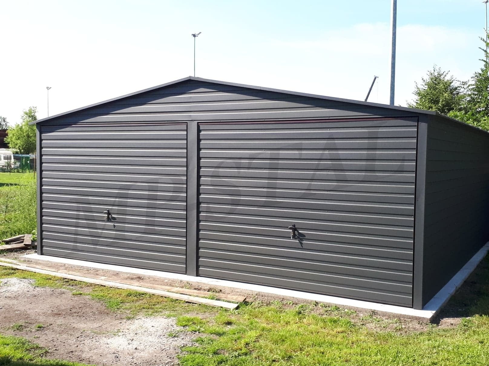 Szary garaż blaszany dwustanowiskowy z dwuspadowym dachem, widoczny z przodu na tle zieleni i słupów oświetleniowych. Garaż posadowiony na betonowej wylewce.