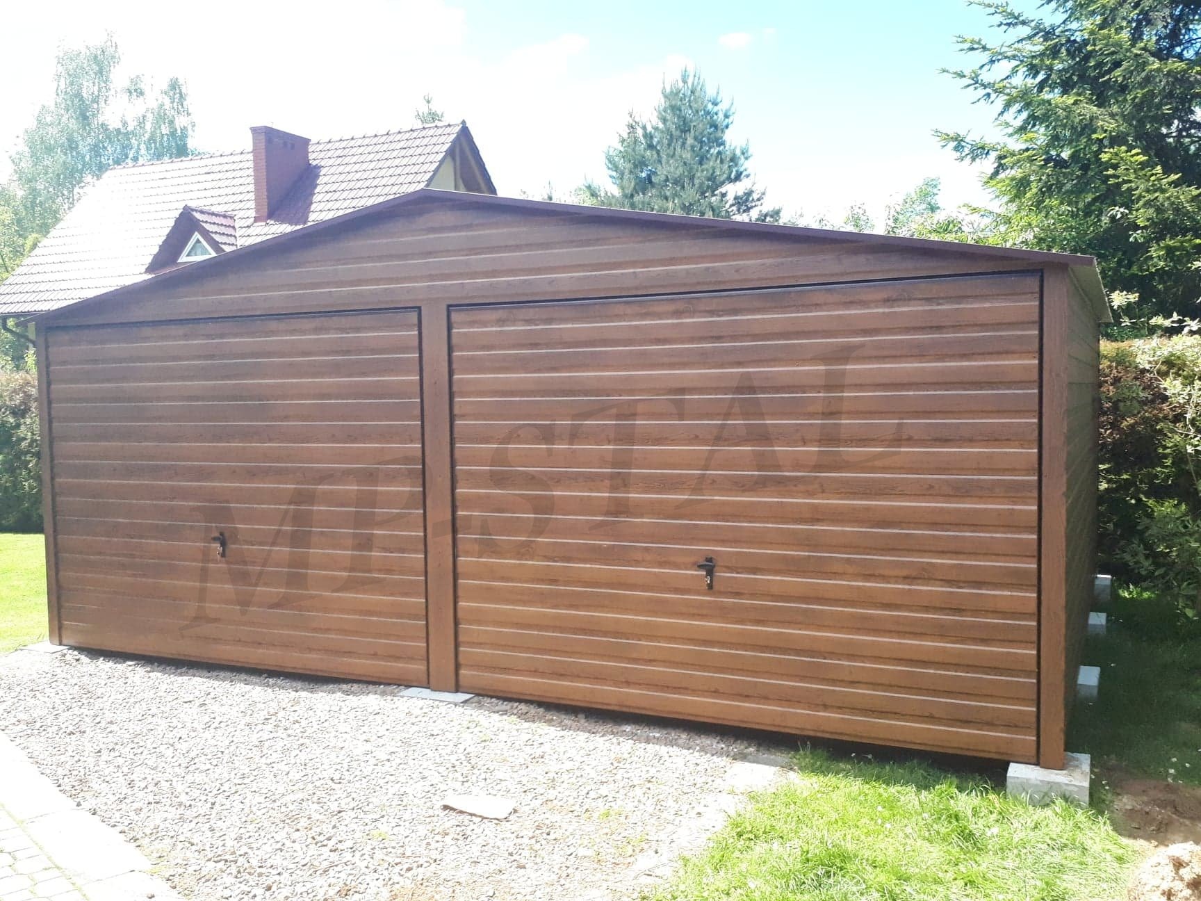 Dwustanowiskowy garaż blaszany z bramami w kolorze drewna, usytuowany na żwirowym podłożu, z domem w tle i zielenią drzew, w słoneczny dzień.