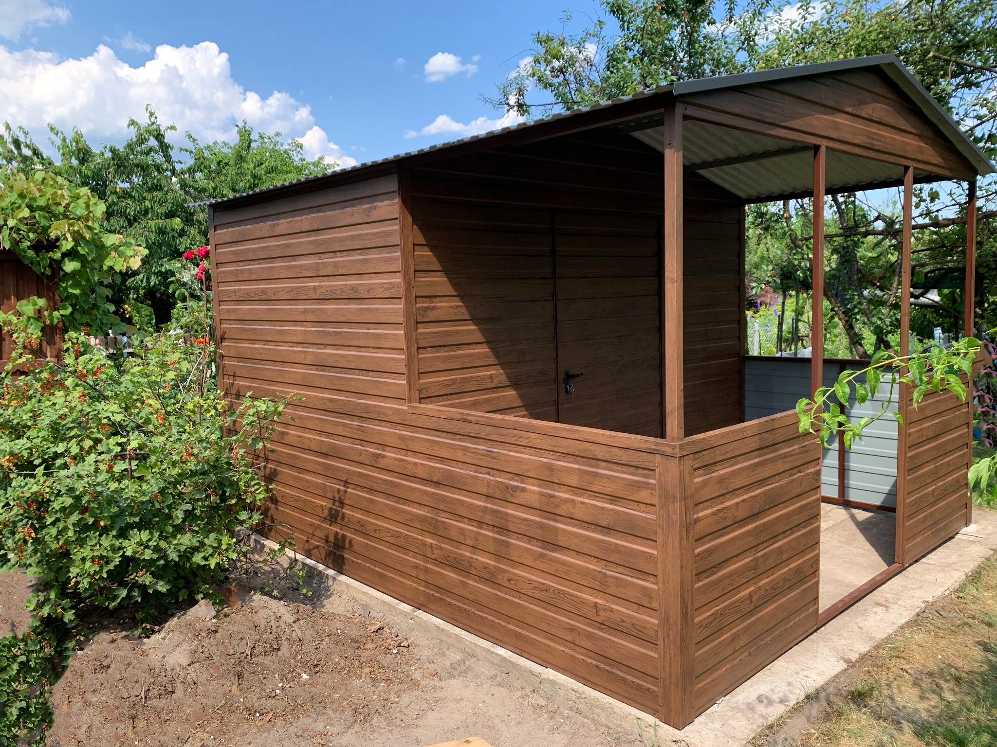 Brązowy garaż blaszany z zadaszeniem, imitujący drewno, otoczony zielenią. Widoczne panele poziome i konstrukcja szkieletowa zadaszenia.