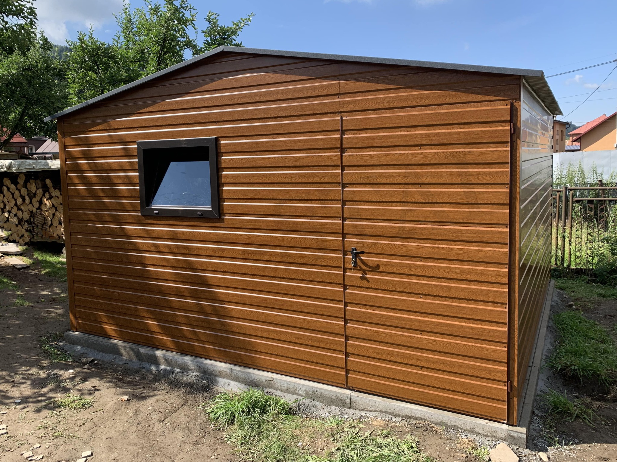 Brązowy garaż blaszany z oknem i drzwiami na betonowej podmurówce, w tle drzewa i domy. Blacha imitująca drewno, poziomy układ paneli, czarna stolarka okienna.