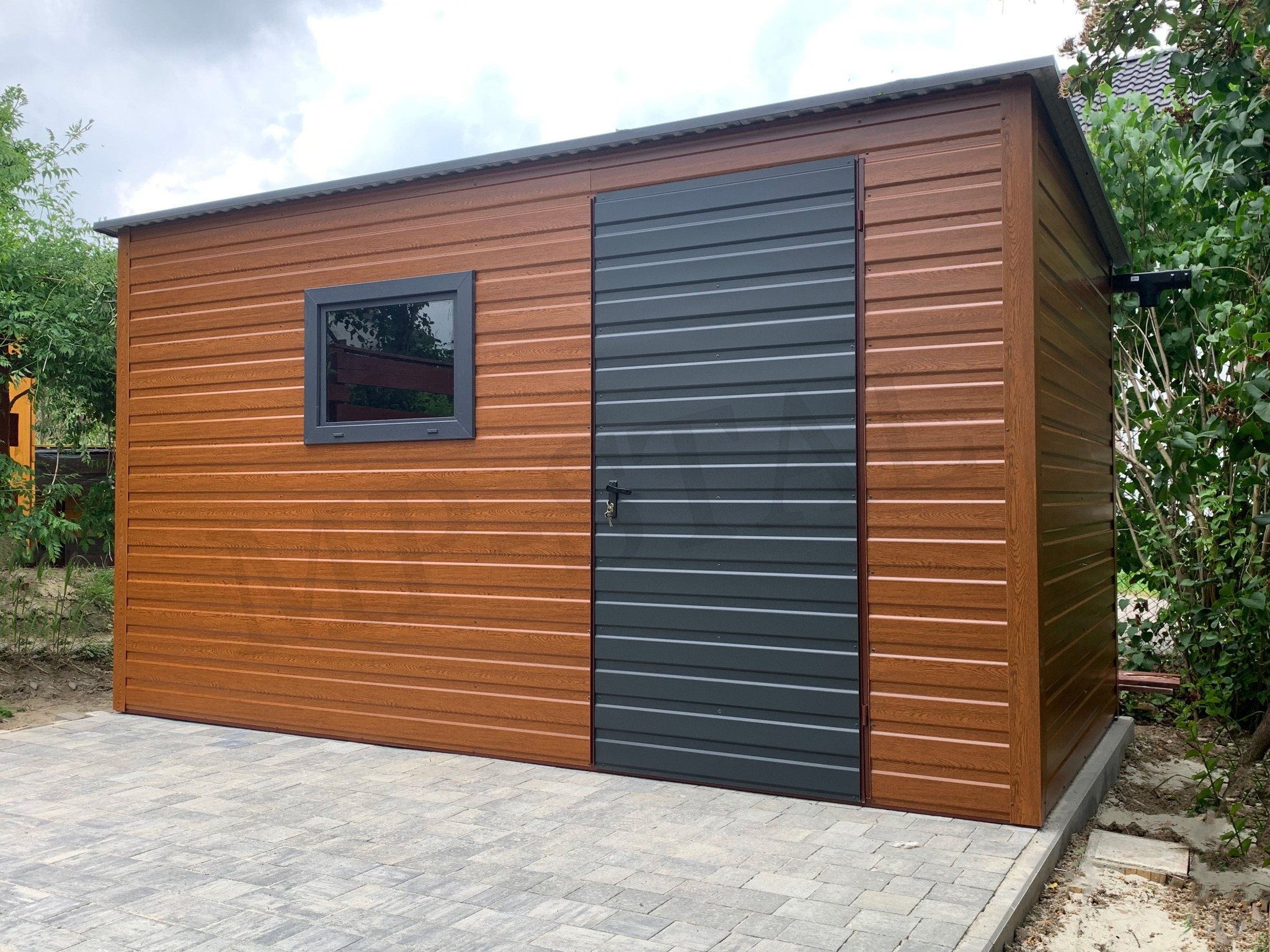 Brązowy garaż blaszany z szarą bramą i oknem na kostce brukowej. Widoczne drzewa i pochmurne niebo.