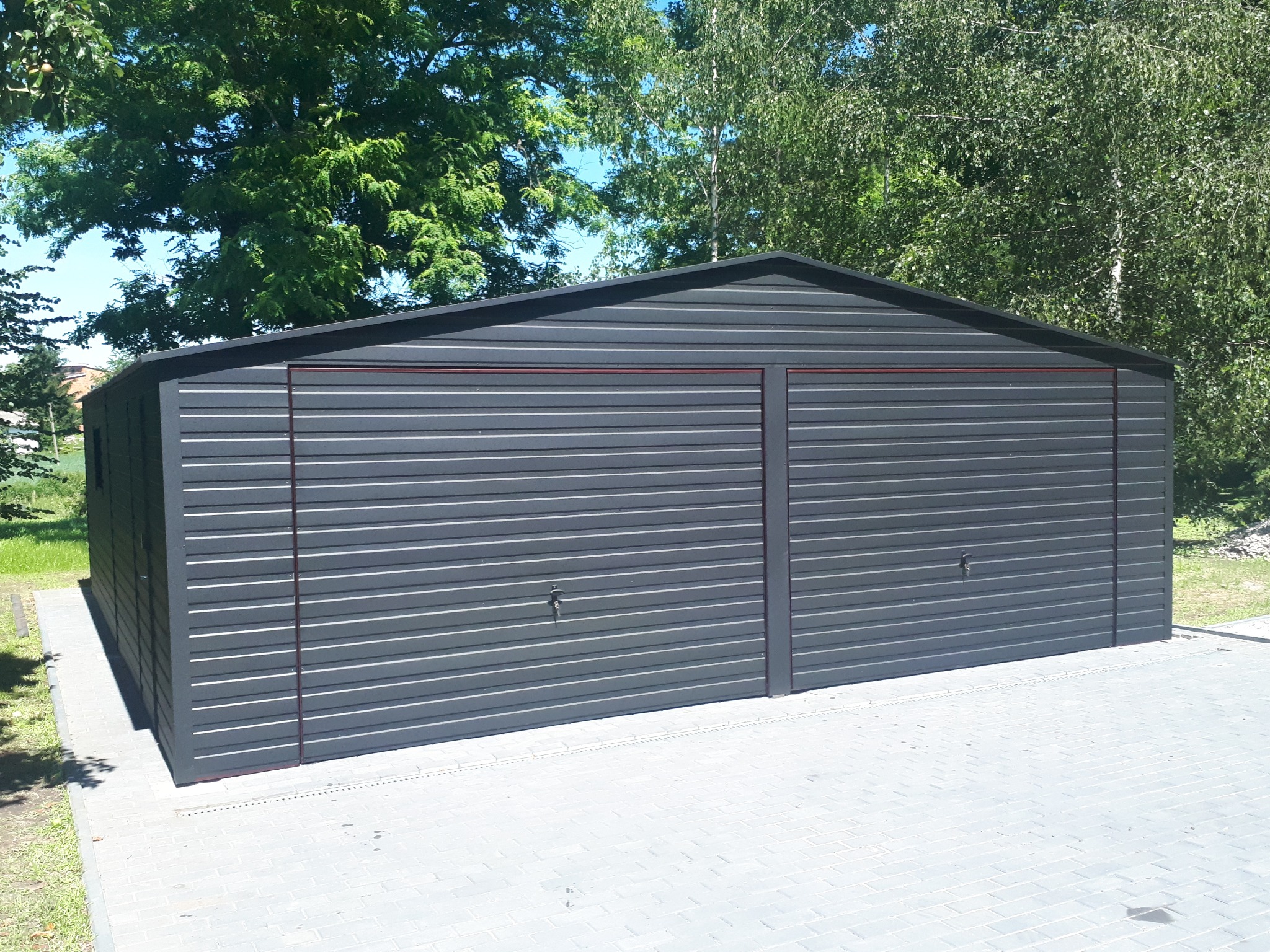 Szary garaż blaszany dwustanowiskowy z dwiema bramami segmentowymi, na kostce brukowej, w tle zieleń drzew. Nowoczesny design, poziome przetłoczenia.