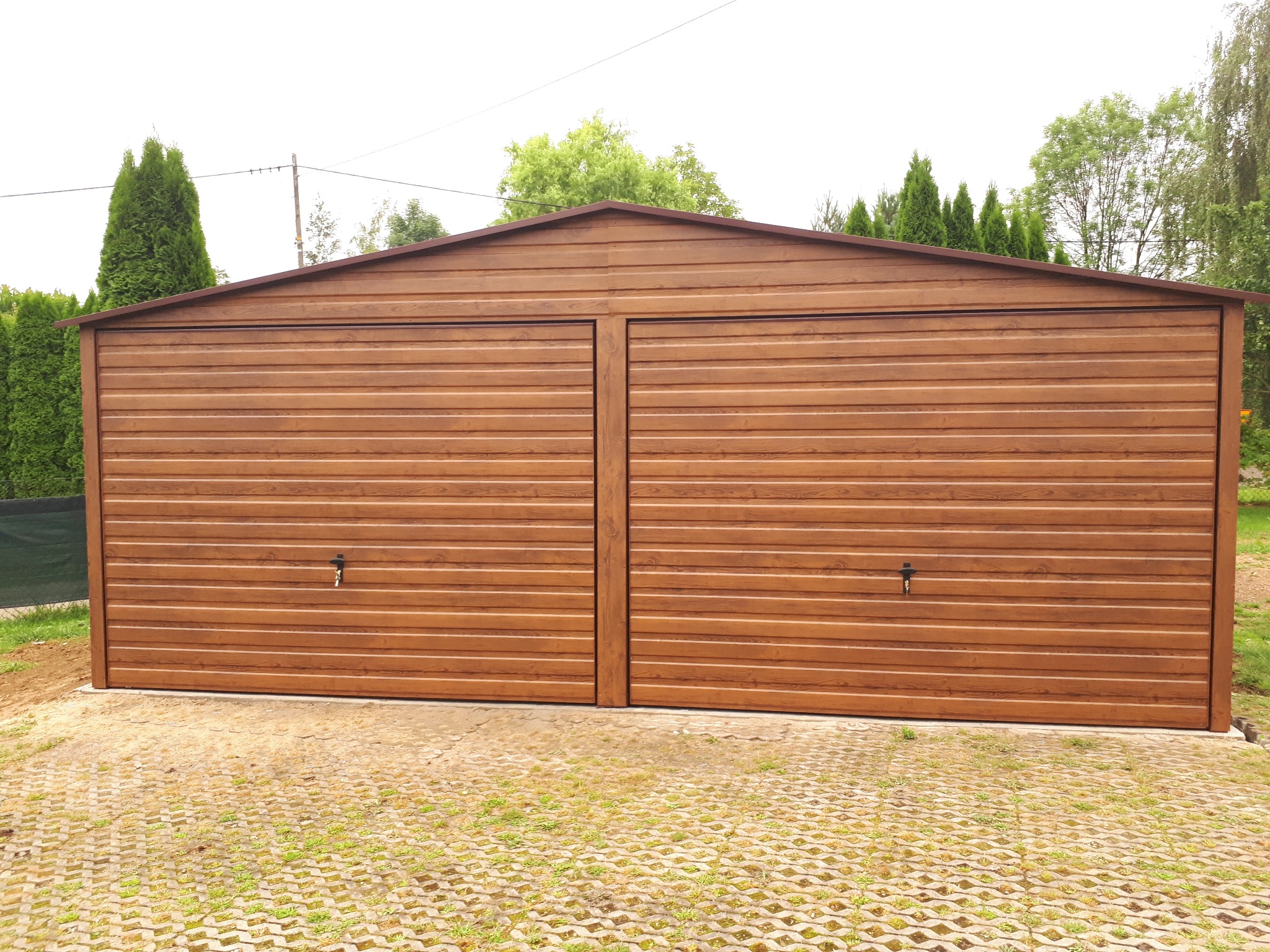 Dwustanowiskowy garaż blaszany w odcieniach brązu, z poziomym wzorem desek, na podłożu z ażurowych płyt betonowych.