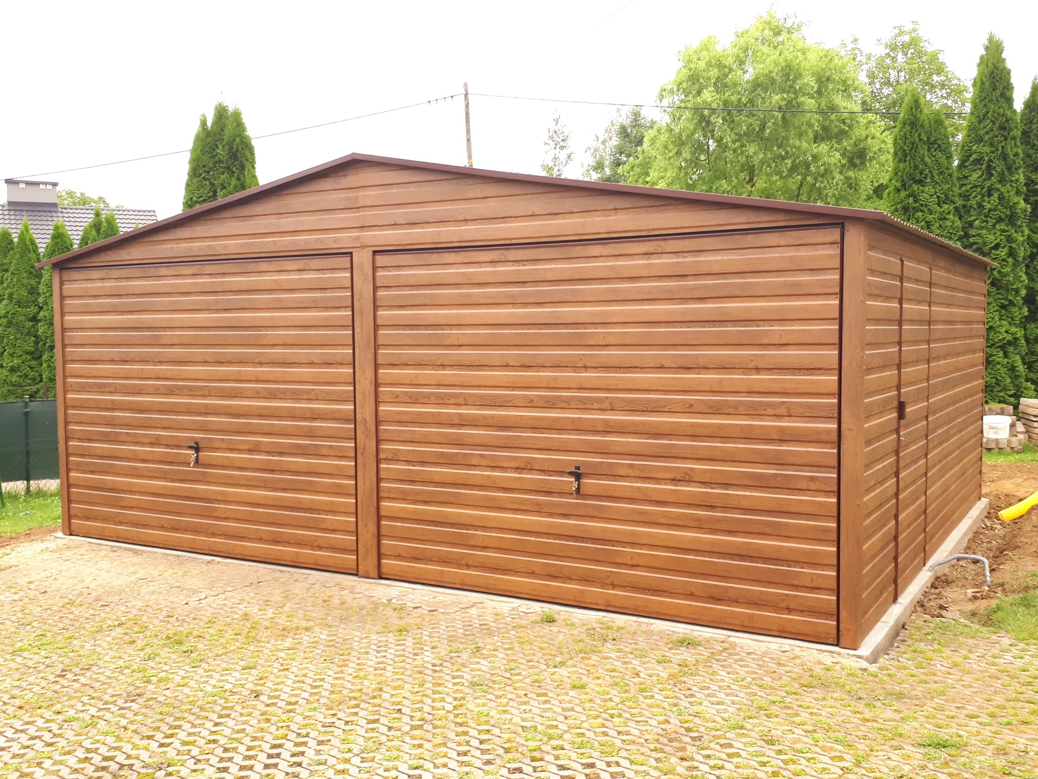 Dwustanowiskowy garaż blaszany w kolorze drewna, z dwuspadowym dachem, na kostce brukowej. Widoczne drzewa w tle i elementy wykończeniowe.