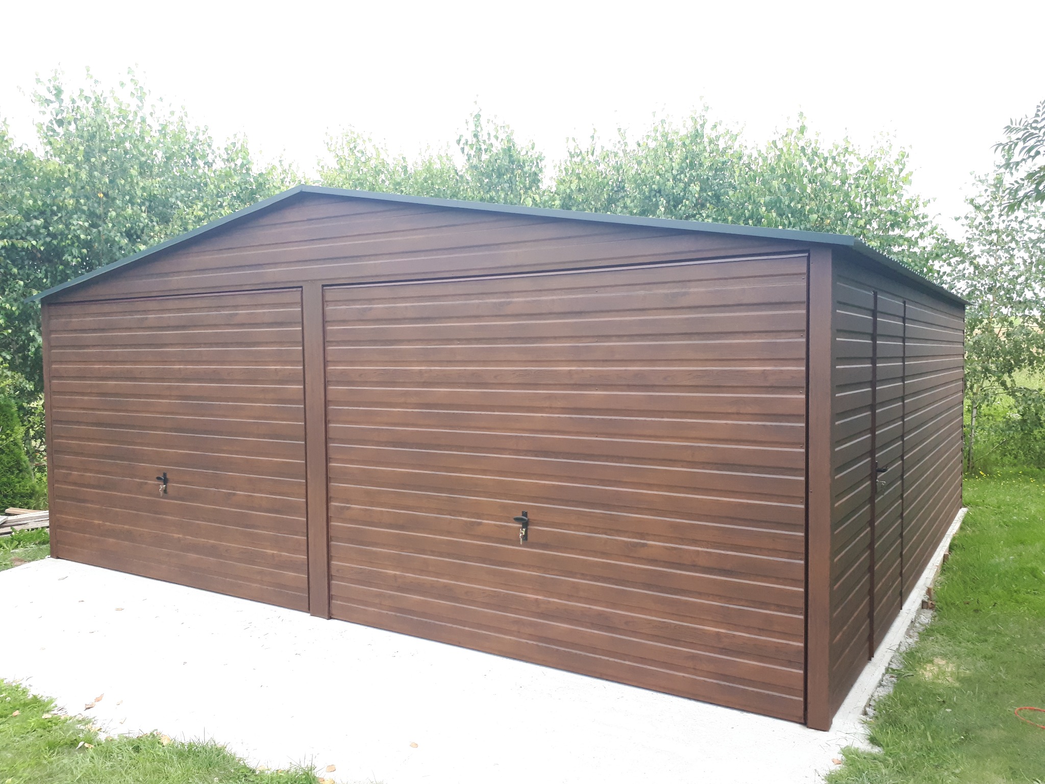 Nowoczesny, brązowy garaż blaszany z dwiema bramami uchylnymi, na betonowej wylewce, otoczony zielenią. Widok z boku, lekko pod kątem, w naturalnym oświetleniu.