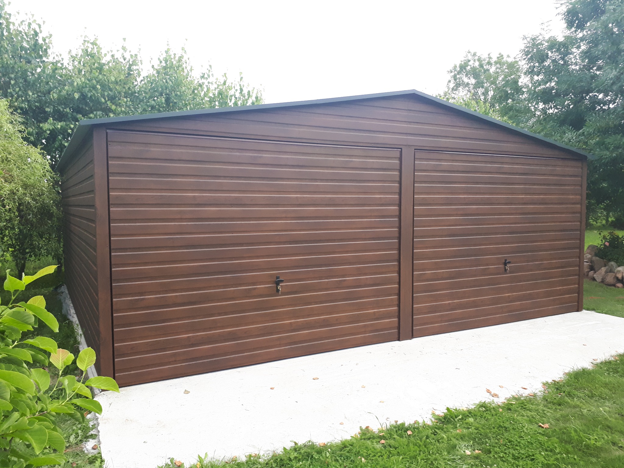 Dwustanowiskowy, brązowy garaż blaszany z poziomym wzorem paneli, na betonowej wylewce, otoczony zielenią. Ujęcie z lekkiego dołu.