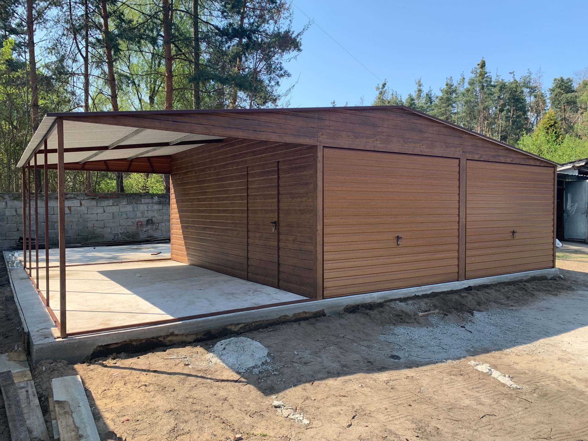 Brązowy garaż blaszany z wiatą na betonowej wylewce, widok z zewnątrz. Na pierwszym planie ziemia, w tle drzewa i fragment betonowego muru.